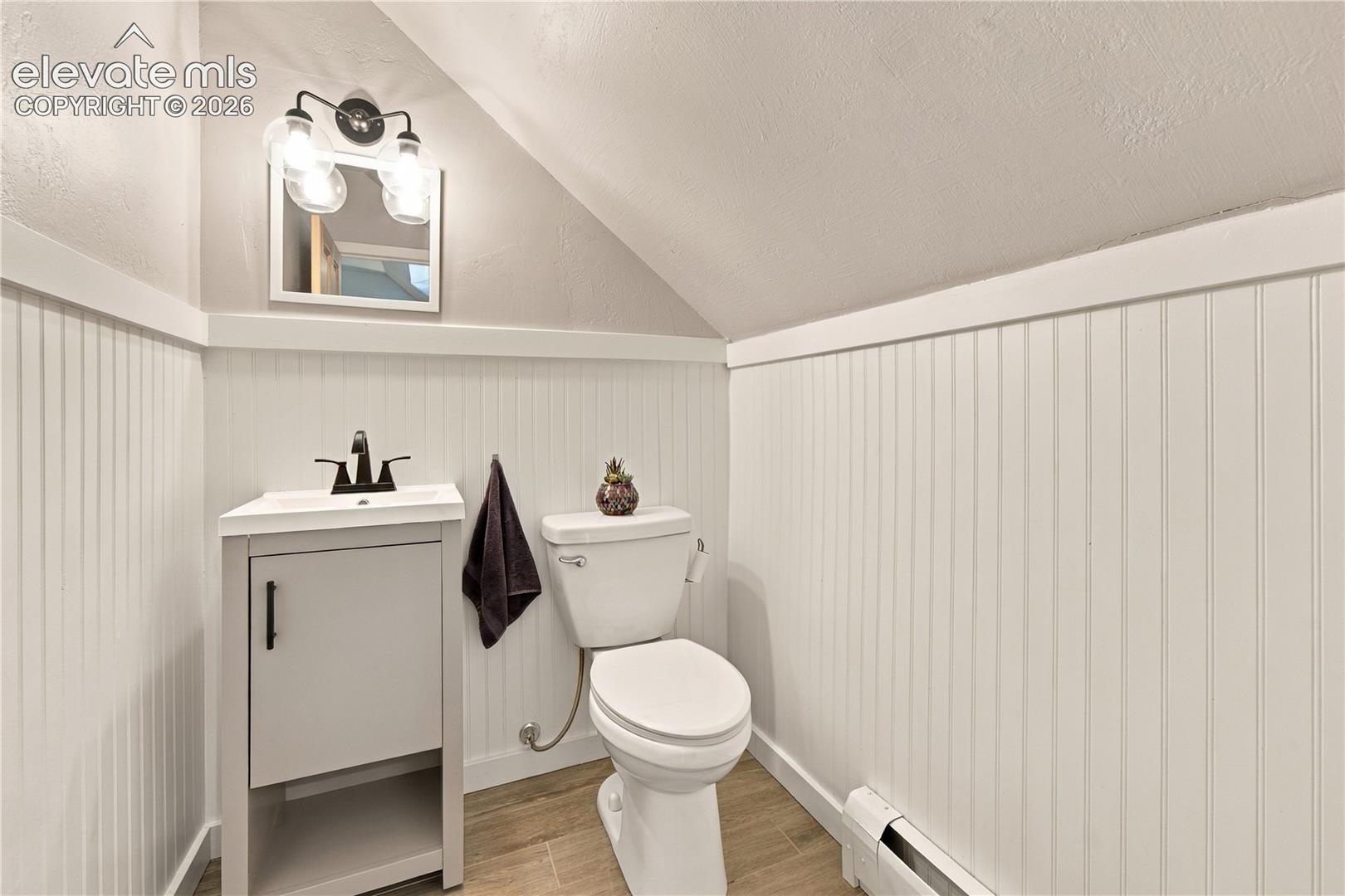 Bathroom featuring white beadboard wainscoting, wood-finish flooring, a neutral-toned vanity with a rectangular sink, matte black hardware, and three-light wall-mounted fixture