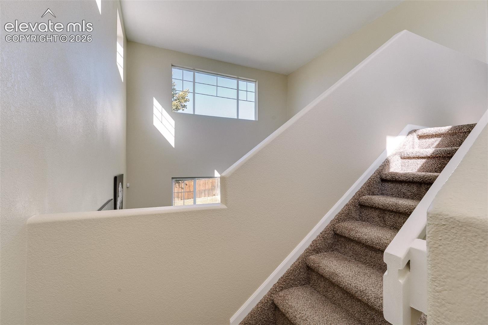 High-ceiling interior with carpeted staircase, wall-mounted white railing, and multiple multi-pane windows