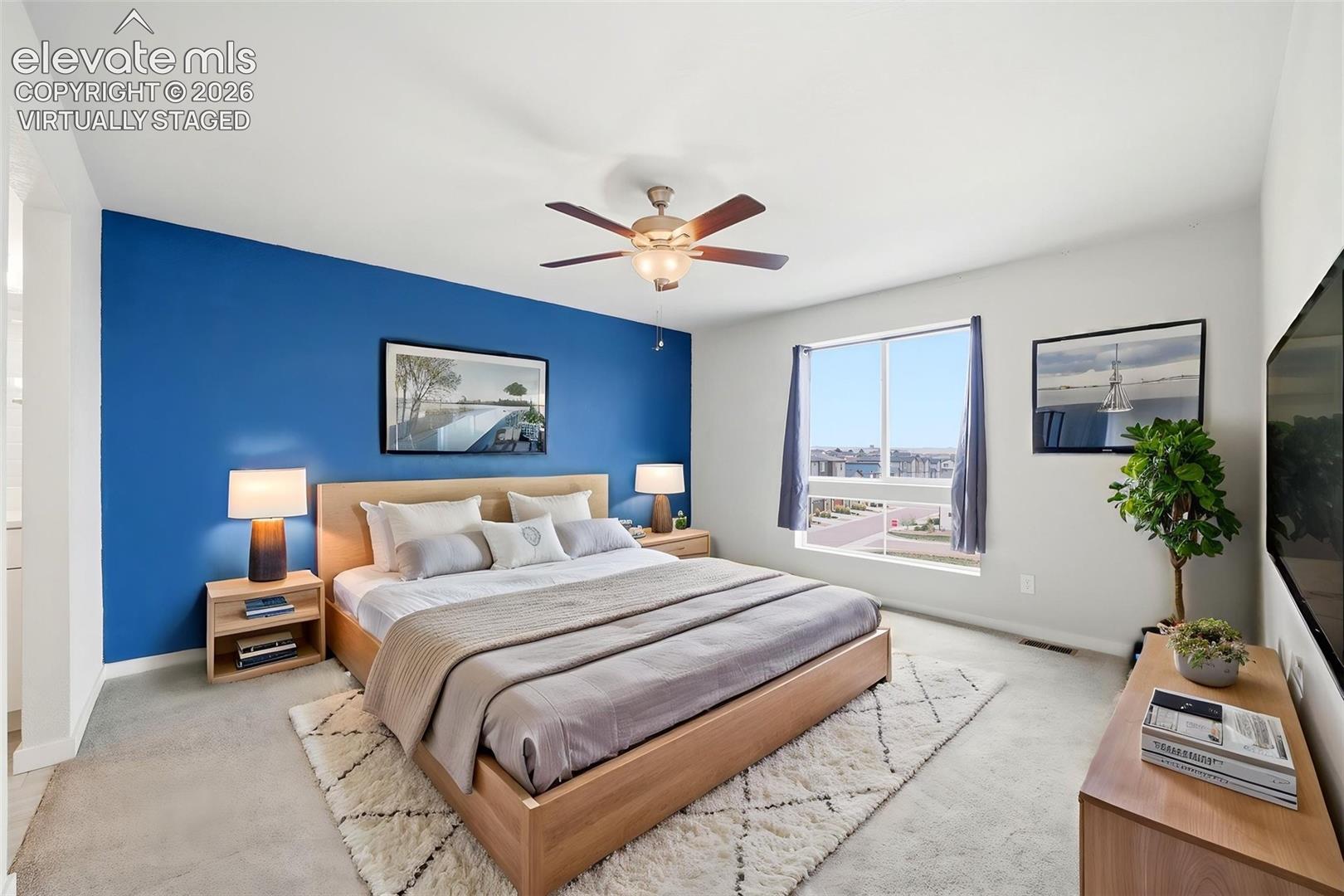 Virtually Staged - Spacious primary bedroom suite featuring an accent wall in rich cobalt blue, wood-frame bed with upholstered headboard, dual nightstands with integrated lighting, neutral carpet flooring, and large sliding glass doors opening to expansive natural light