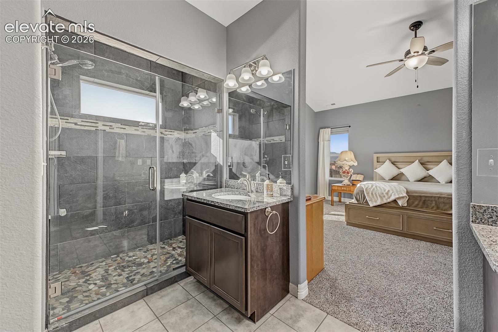 Walk-in shower with dark tile surround and stone flooring, stone-topped dual vanities vanity with undermount sinks and a walk-in closet. 