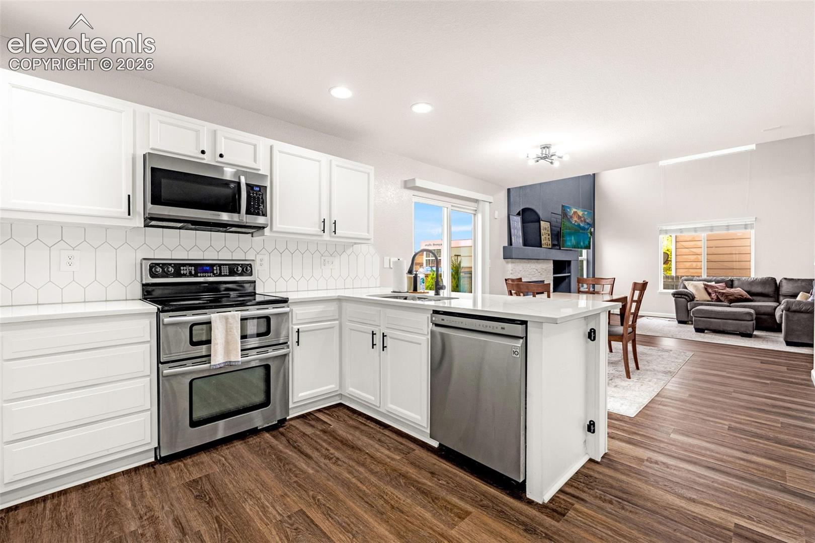 Kitchen featuring white shaker-style cabinetry, stainless steel appliances, hexagonal tile backsplash, stone-surface countertops, and wood-finish flooring