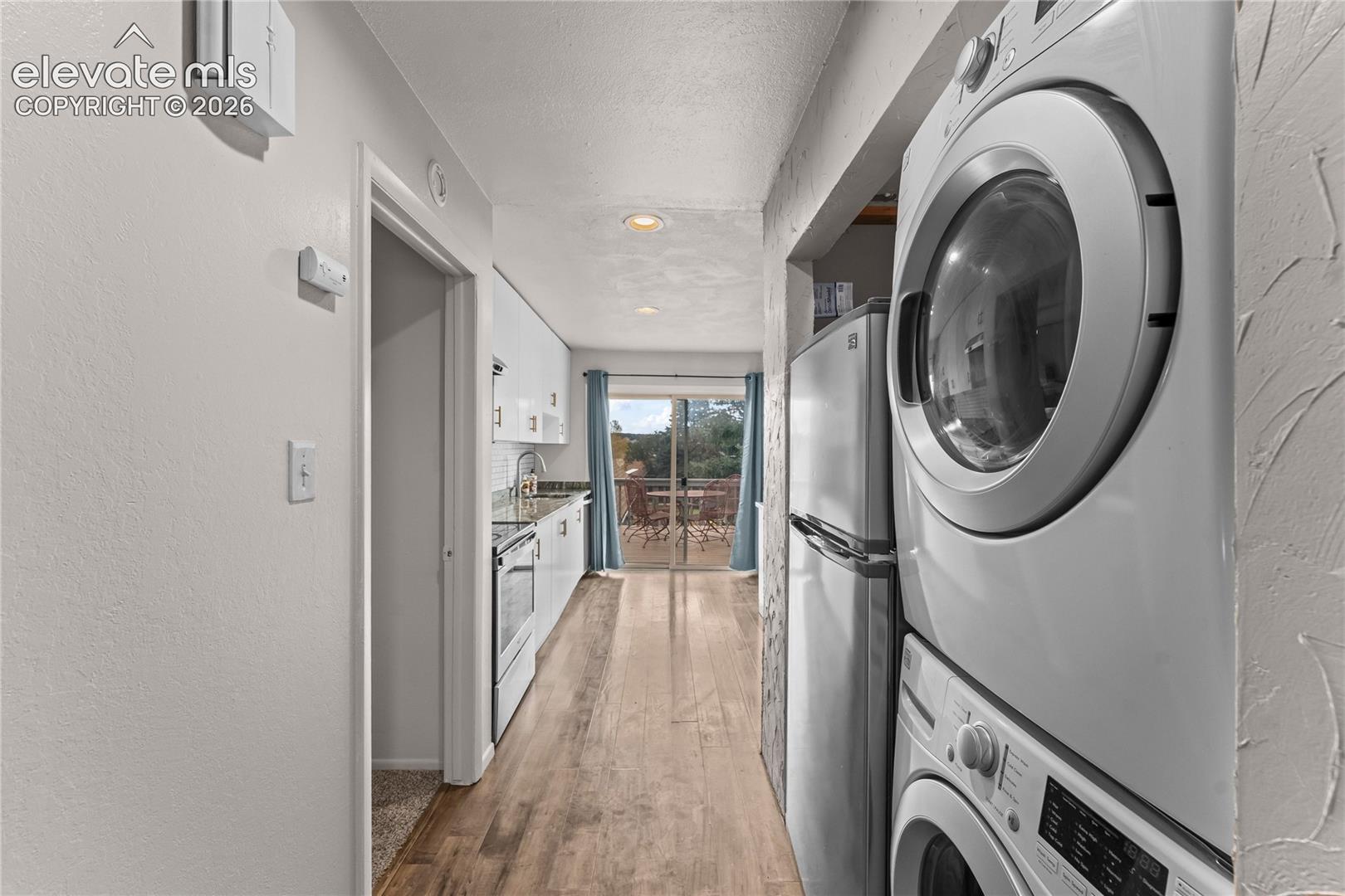 Stacked laundry center, wood-finish flooring, recessed lighting, and kitchen area featuring white cabinetry and stainless steel refrigerator