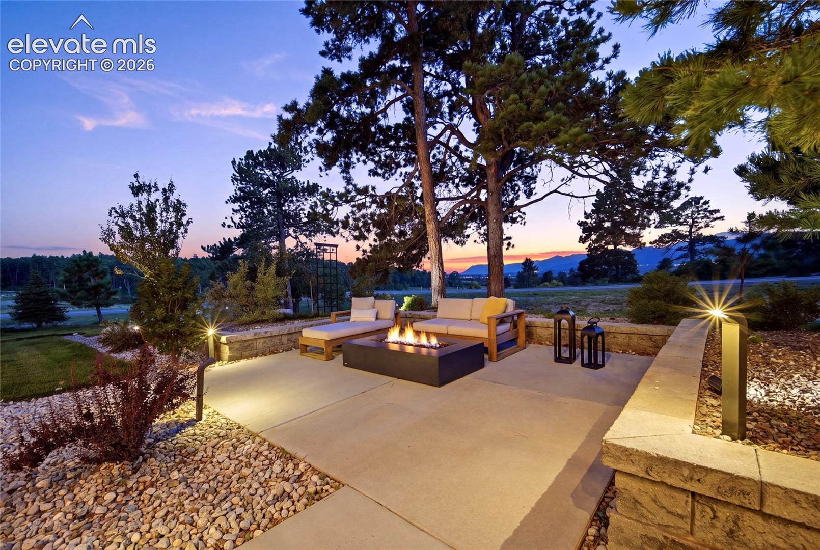 Expansive patio with a built-in fire feature, surrounded by landscaping with path lighting, and a stone retaining wall