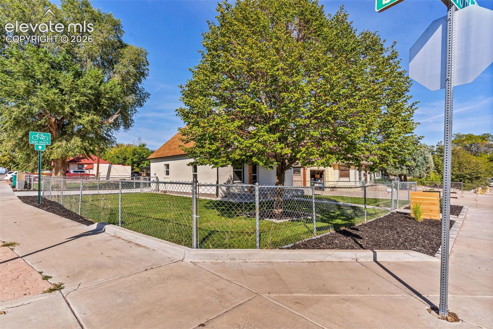 Corner lot property featuring a fenced perimeter, mature tree canopy, manicured lawn, and mulched landscaping beds
