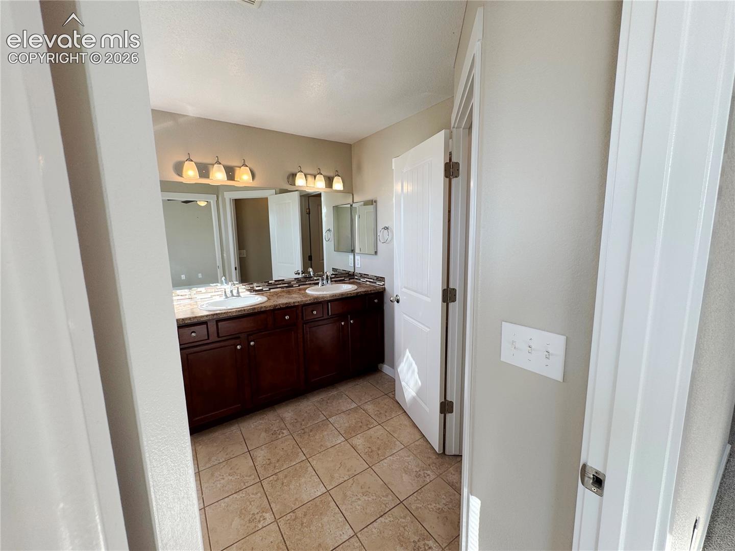 Dual-sink vanity with dark wood cabinetry, stone-finish countertops, tiled flooring, and multi-bulb wall lighting