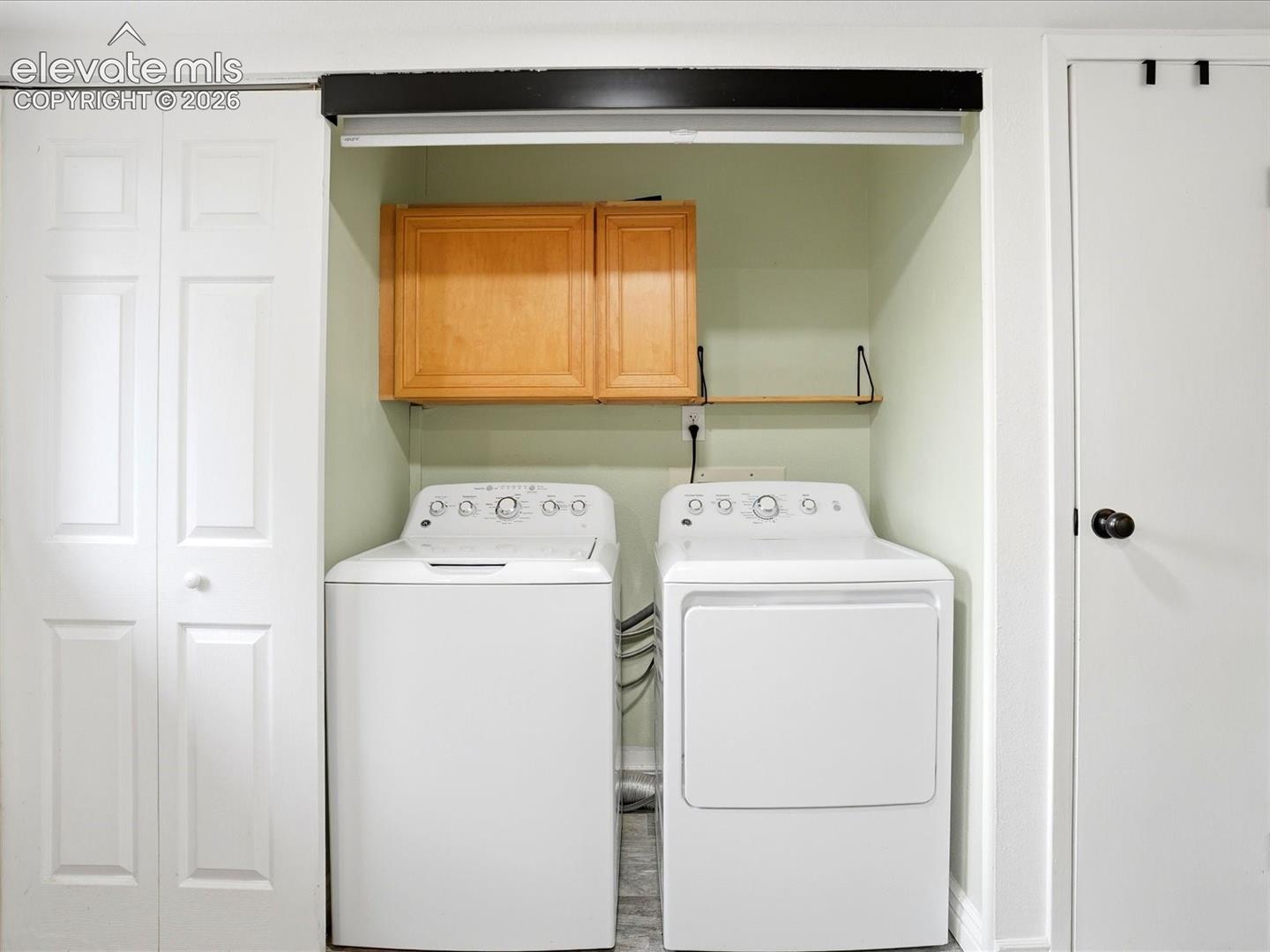 Laundry area featuring wood-finish wall-mounted cabinetry, an open storage shelf, dual bifold closet doors, and a standard door with a matte black knob