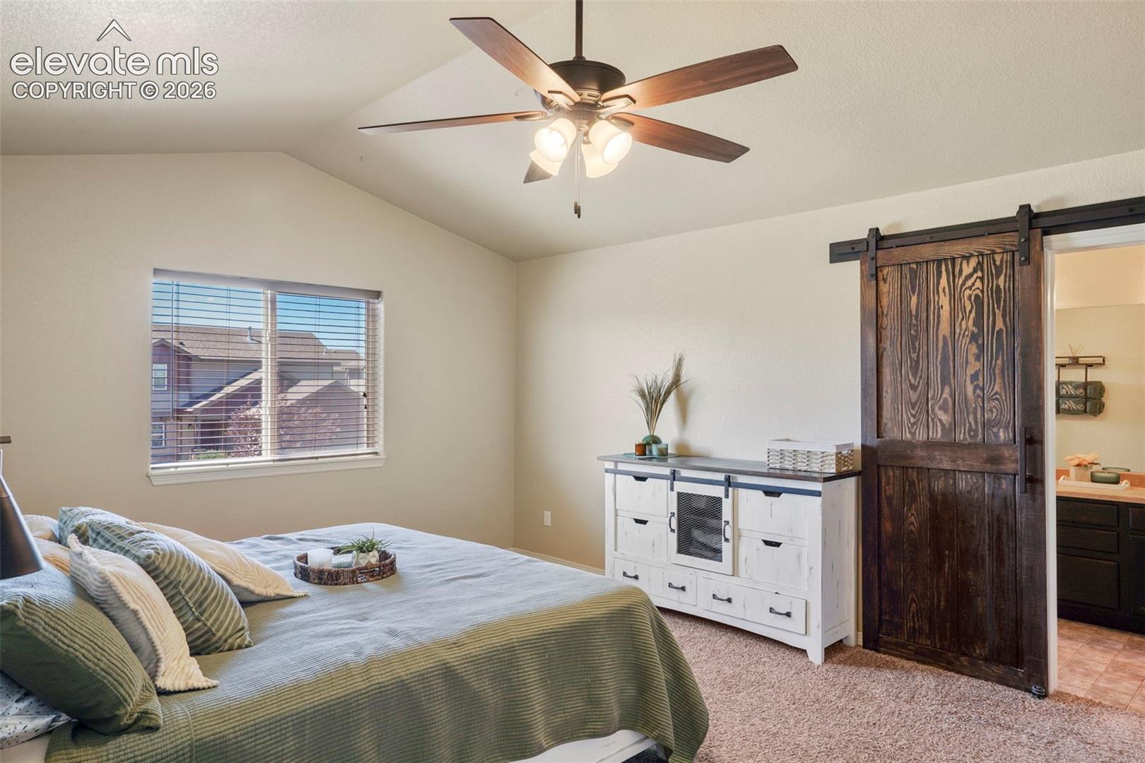 Vaulted ceiling with ceiling fan, window with horizontal blinds, wood-finish sliding barn door, and carpet flooring