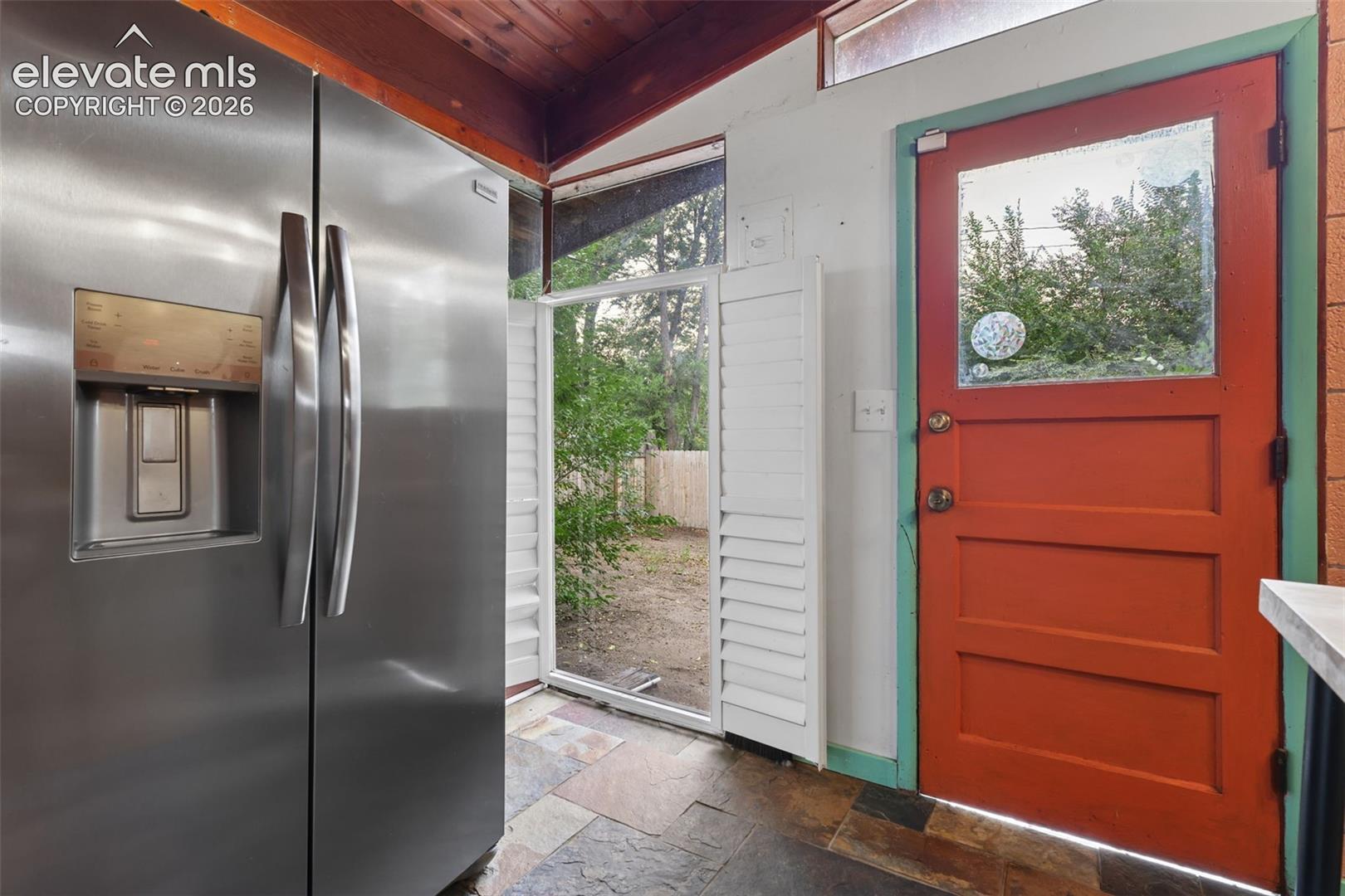 Stainless steel side-by-side refrigerator, slate flooring, wood-paneled ceiling, and a vibrant red panel door with a clear glass insert