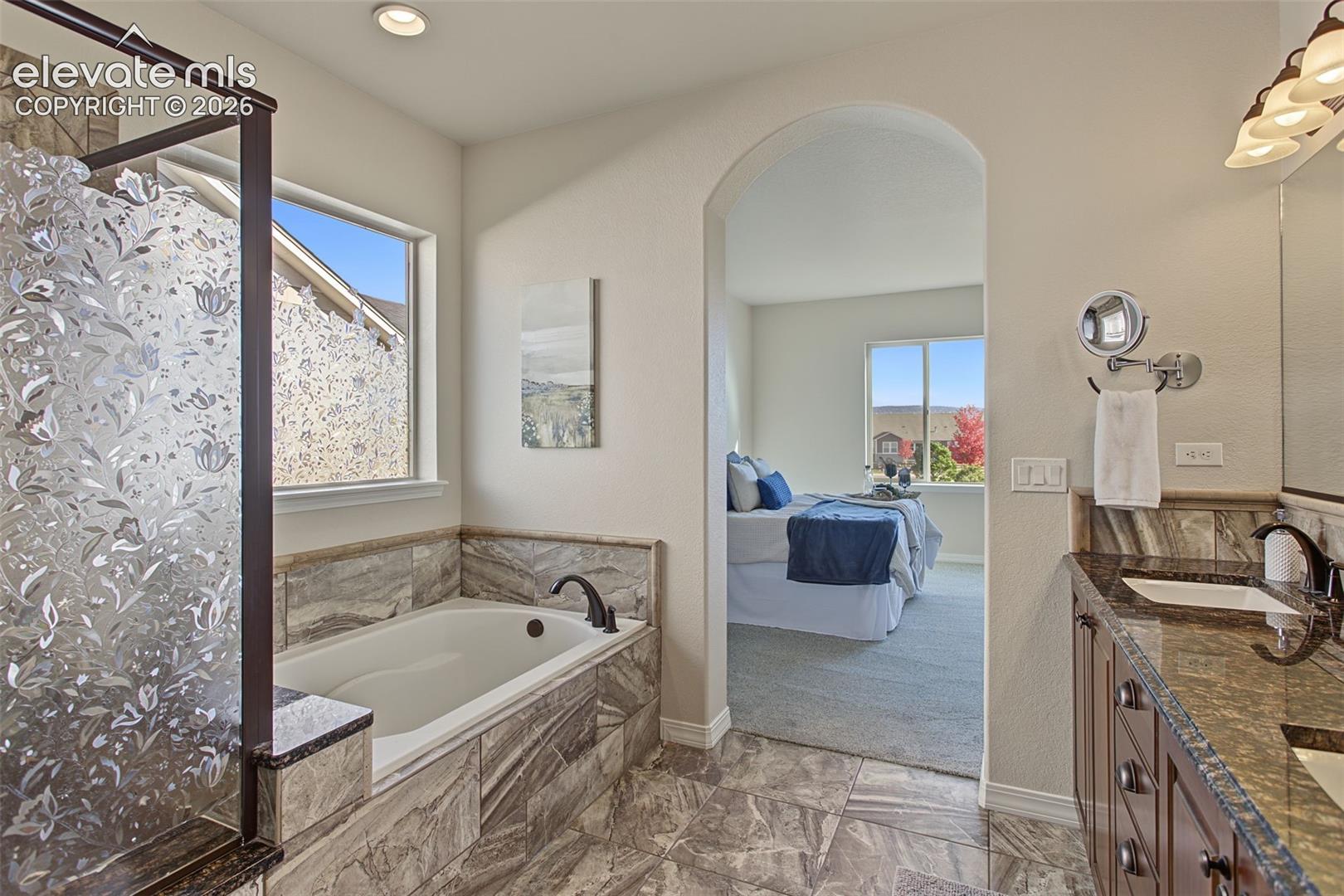 Bathroom featuring a stone-surround soaking tub, dual-sink vanity with stone countertop, walk-in shower with decorative privacy glass, and an arched entry