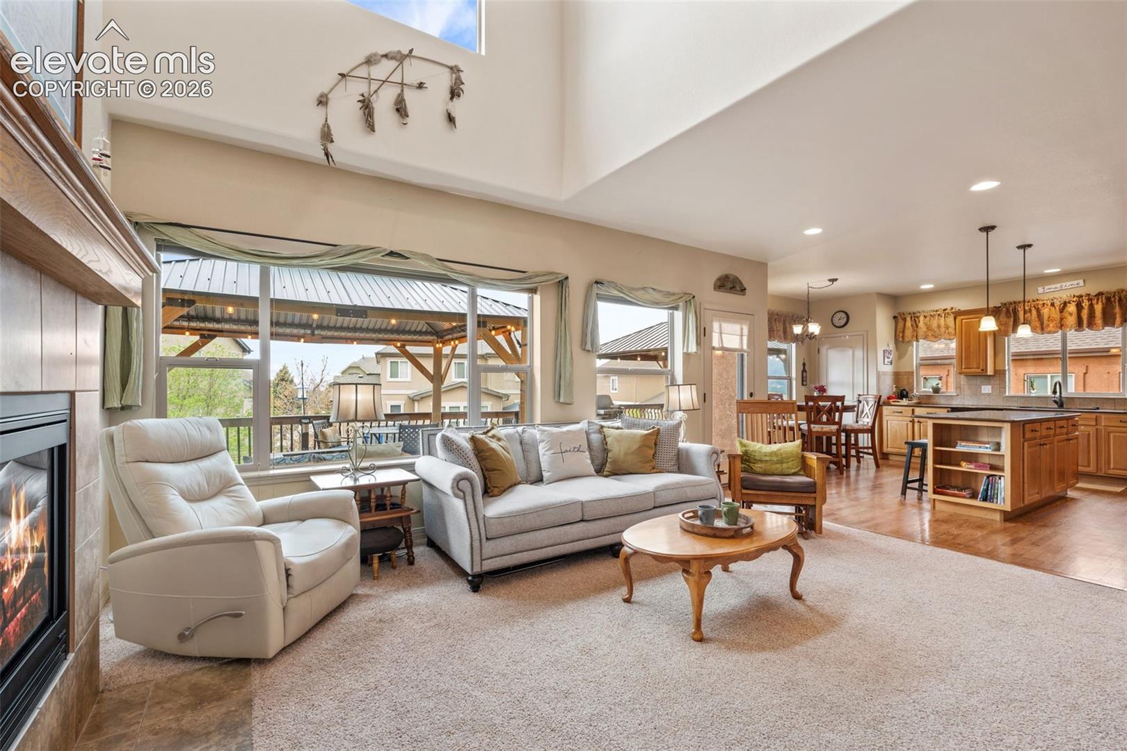 Expansive living area featuring a fireplace with a wood mantle, large windows, vaulted ceilings, and an open-concept design to the kitchen