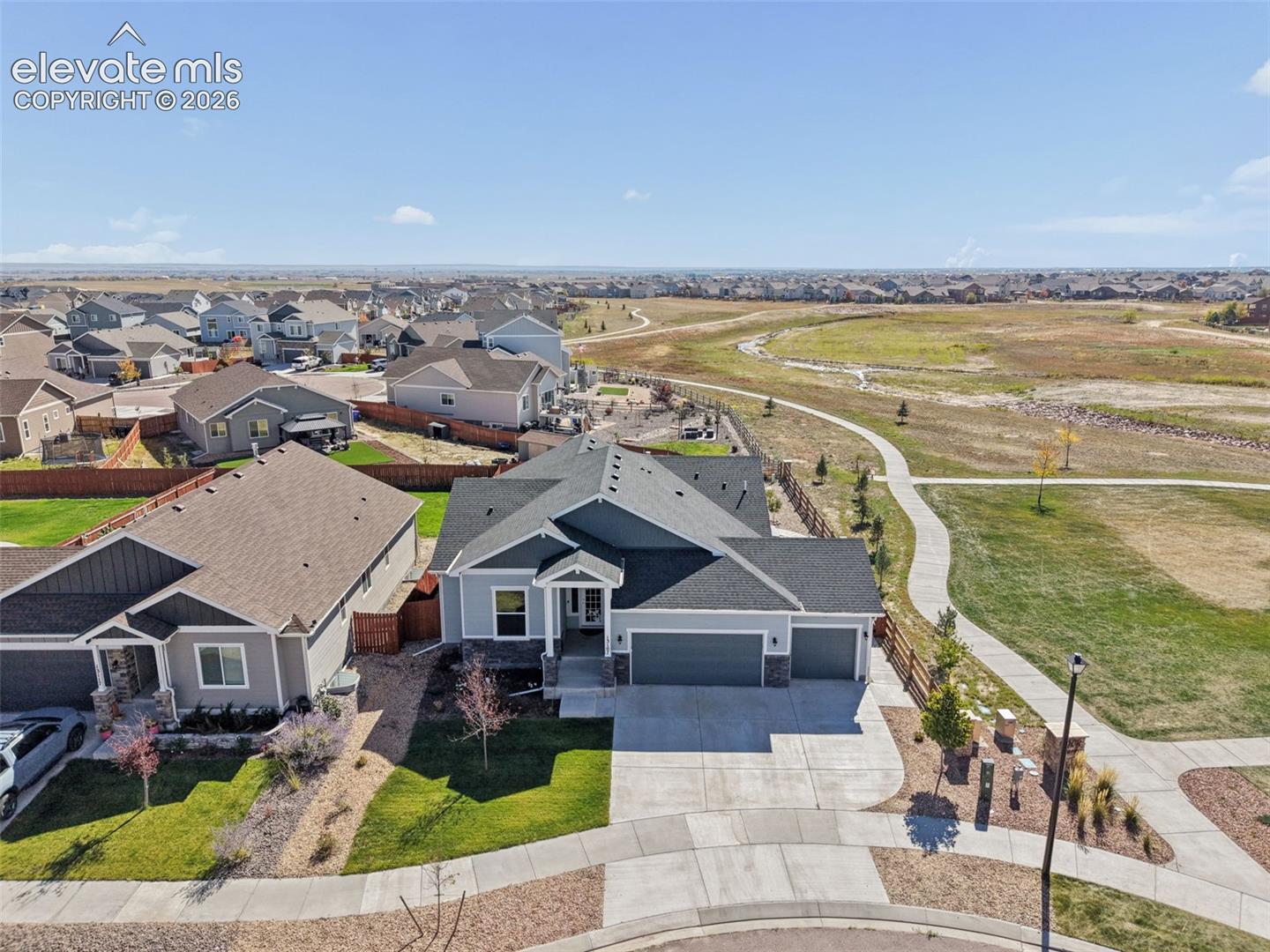 Single-story residence featuring a multi-gabled roofline, stone veneer accents, three-car garage, and concrete driveway, situated on a lot bordering a paved walking path