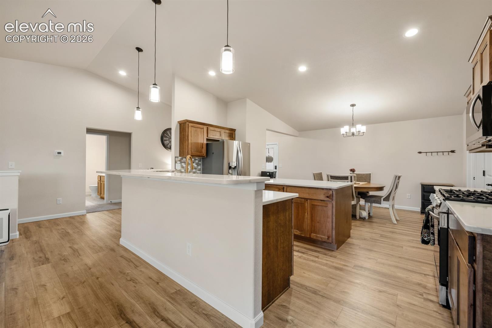 Kitchen featuring a dual-level island with quartz countertops, wood-finish cabinetry, wood-finish flooring, vaulted ceilings, and multi-pendant lighting