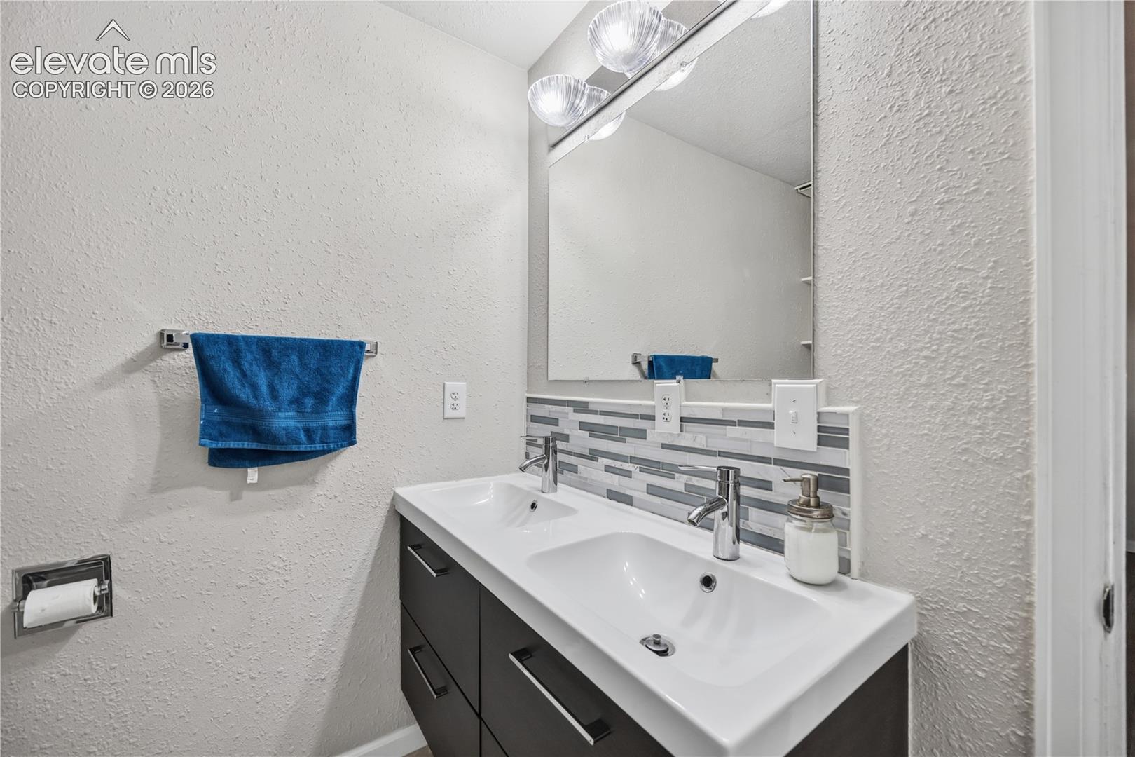 Dual-sink vanity with a solid-surface countertop, linear multi-toned tile backsplash, oversized wall-mounted mirror, and multi-bulb polished chrome vanity lighting