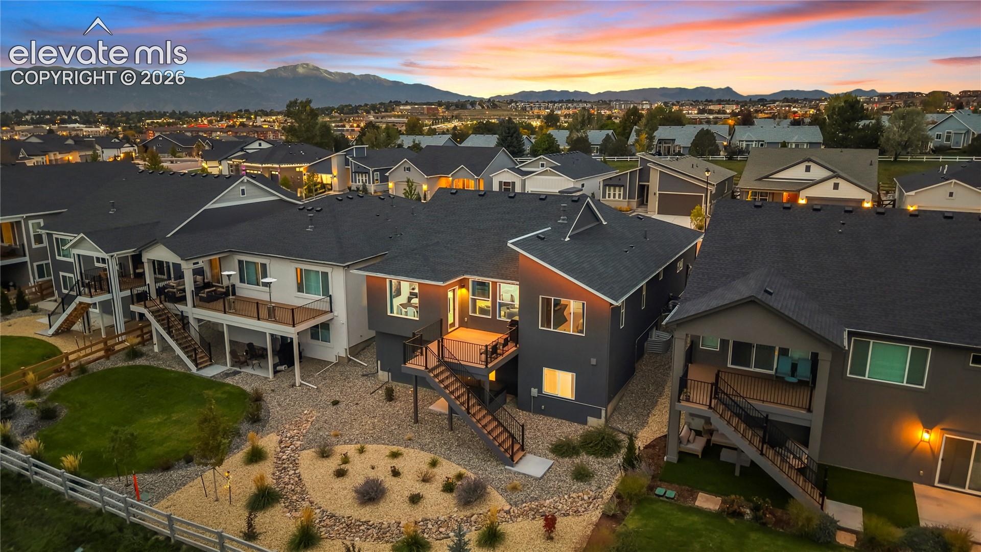 Exterior of multi-story residence featuring composite shingle roofing, elevated wood deck with black metal railing, stairs to lower-level exterior, xeriscaped ground cover, and mountain backdrop
