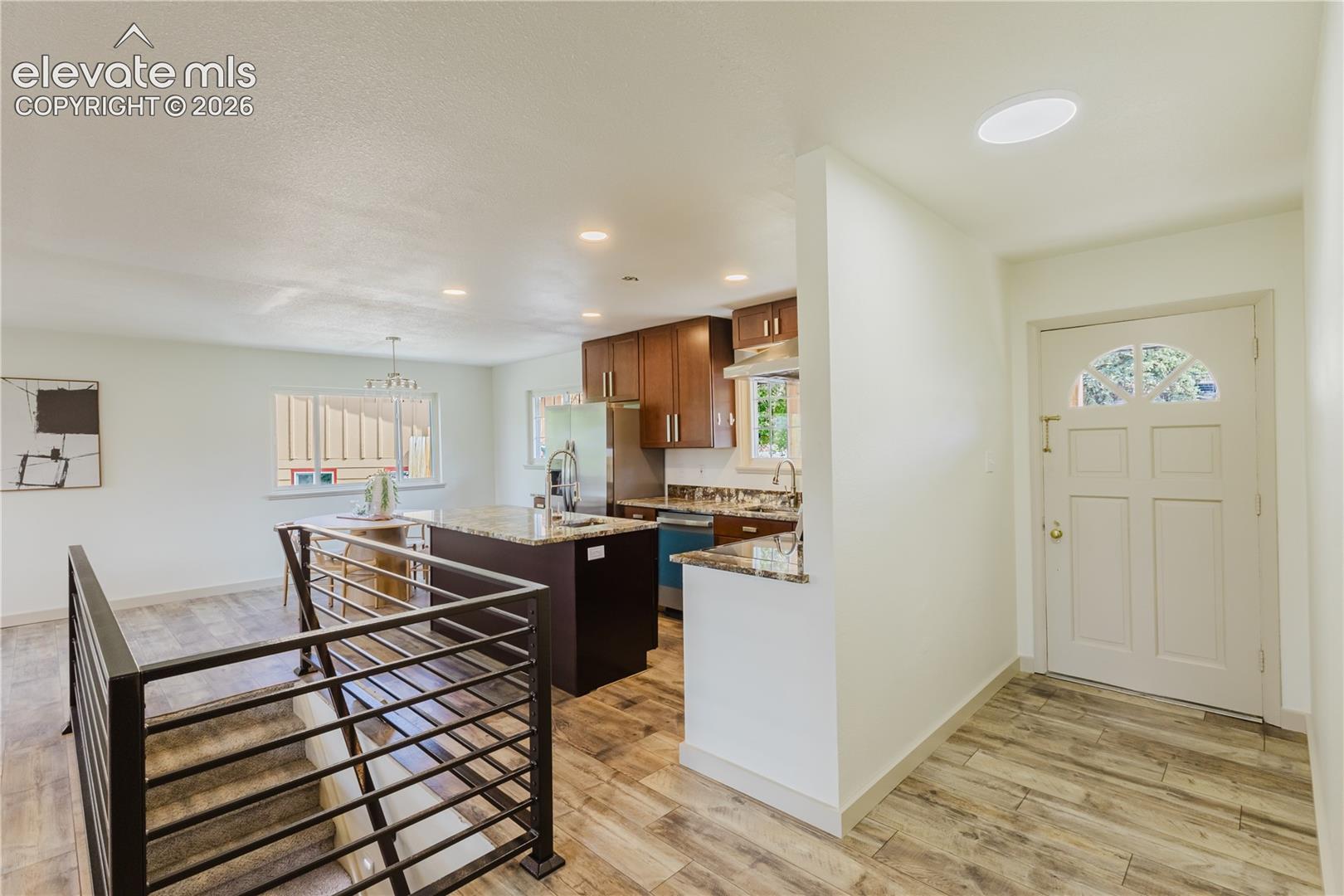 Open-concept living area featuring wood-finish flooring, a contemporary staircase with metal railings, and a kitchen with warm wood cabinetry and granite countertops