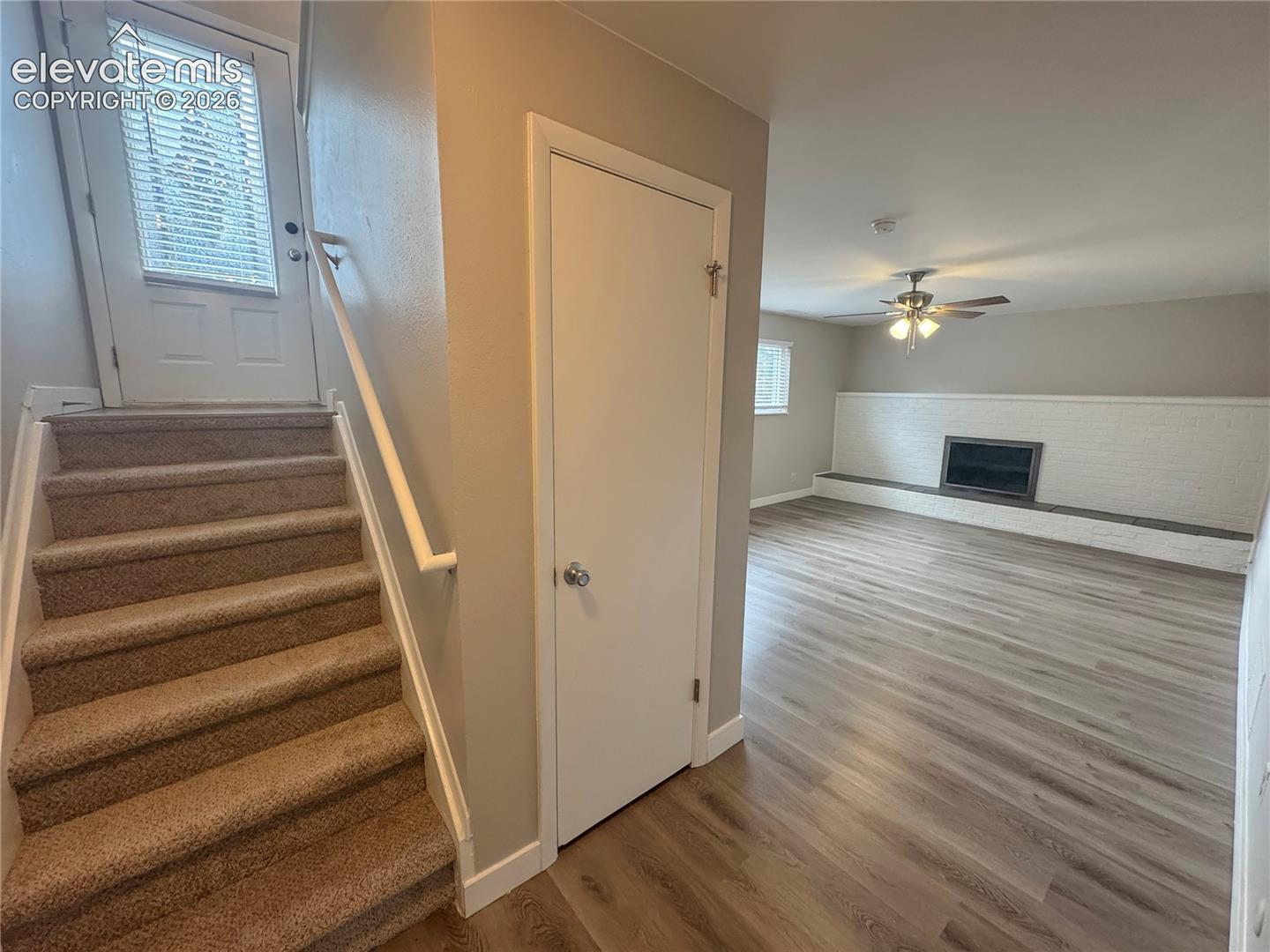 Wood-finish flooring, white painted brick fireplace with hearth, carpeted staircase, and ceiling fan with integrated lighting