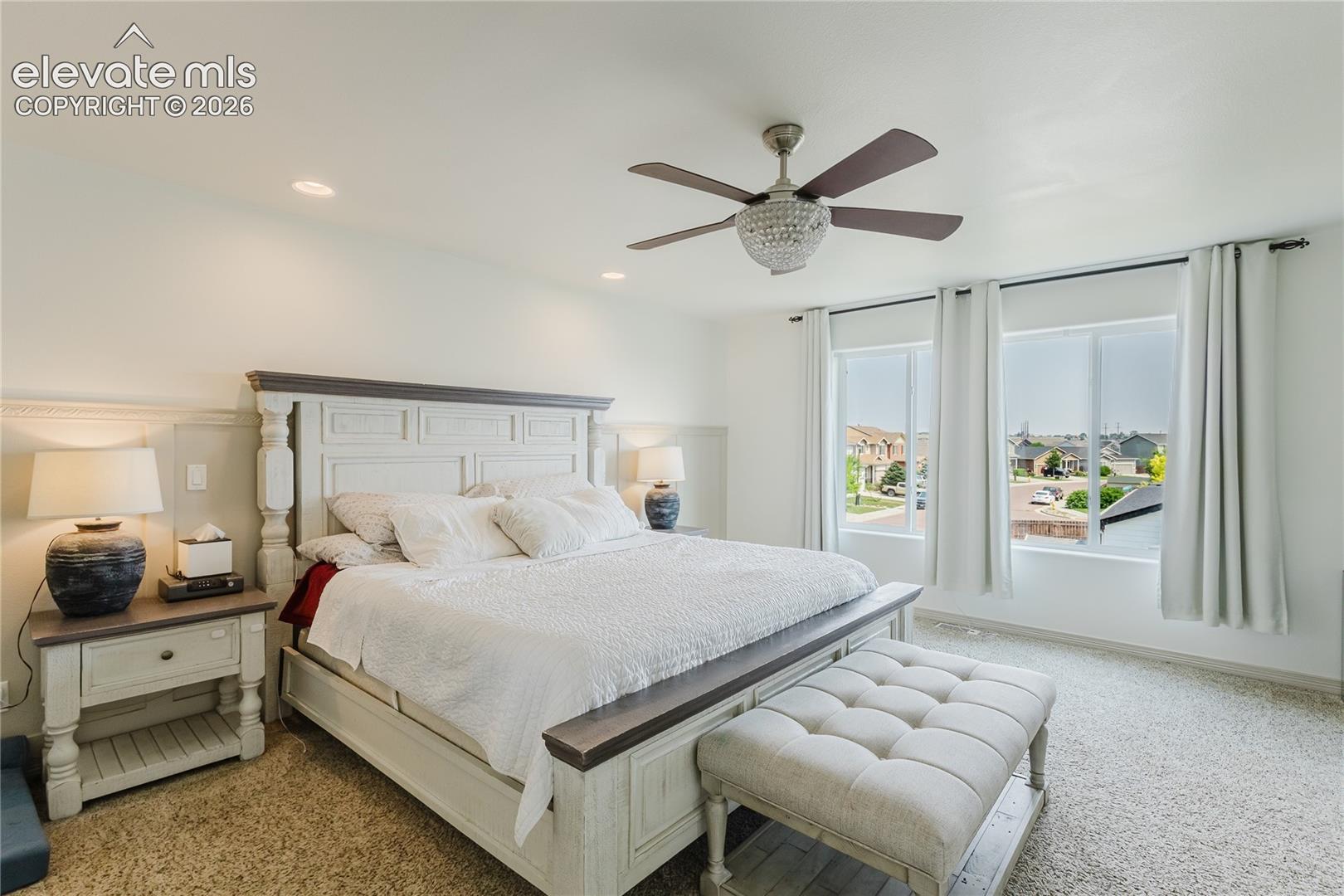 Spacious bedroom featuring light-colored carpet, two large windows, a ceiling fan with integrated lighting, recessed ceiling lights, and white wainscoting