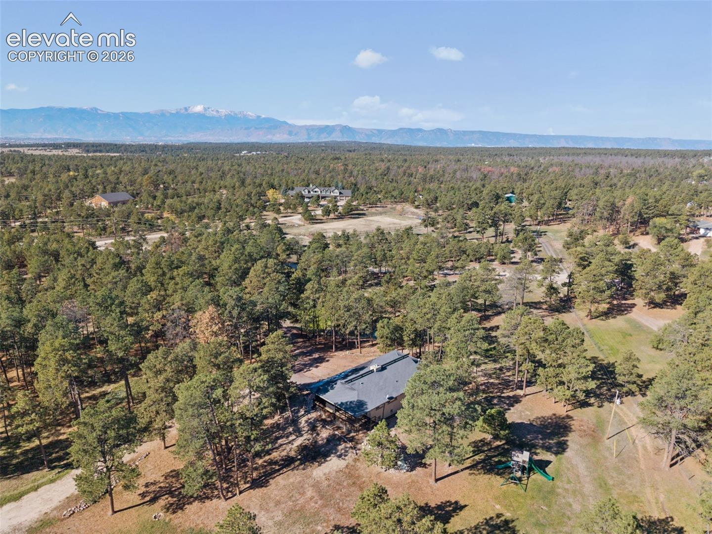 Aerial perspective of a residence nestled within a vast, wooded lot, featuring a metal roof and a backdrop of mountain scenery