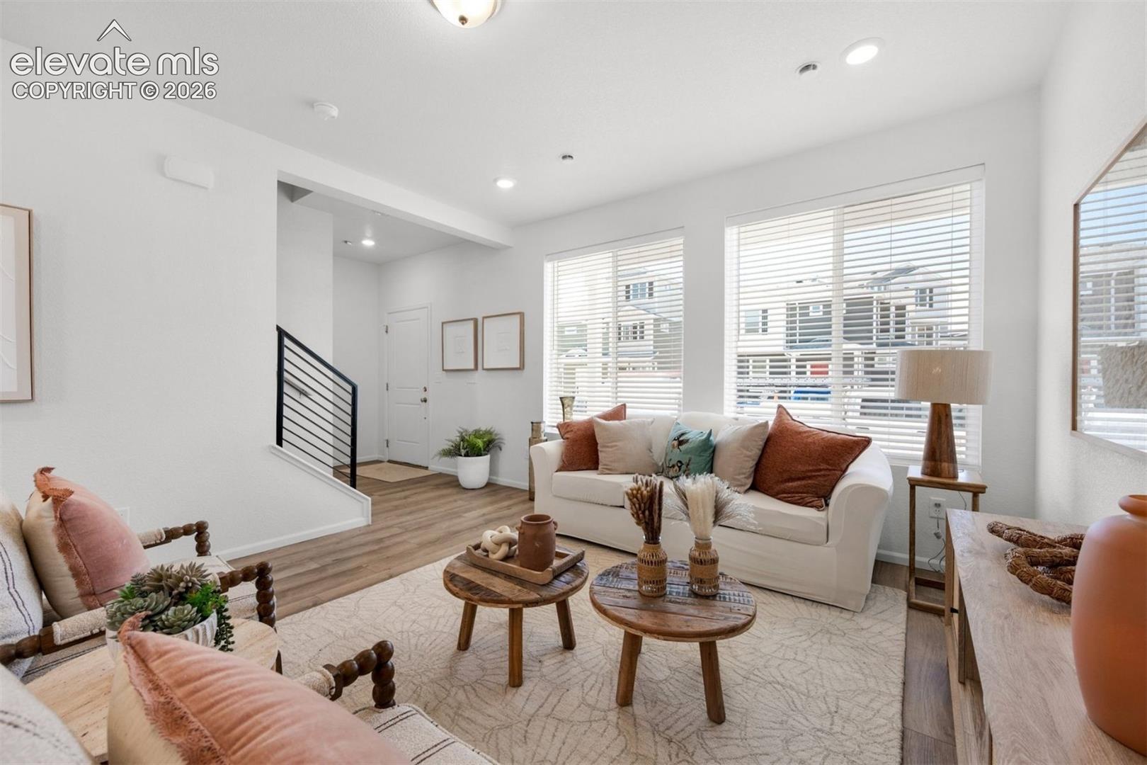 Bright living area featuring wood-finish flooring, multiple windows with blinds, recessed lighting, and a staircase with a black metal railing
*photos of a model home*