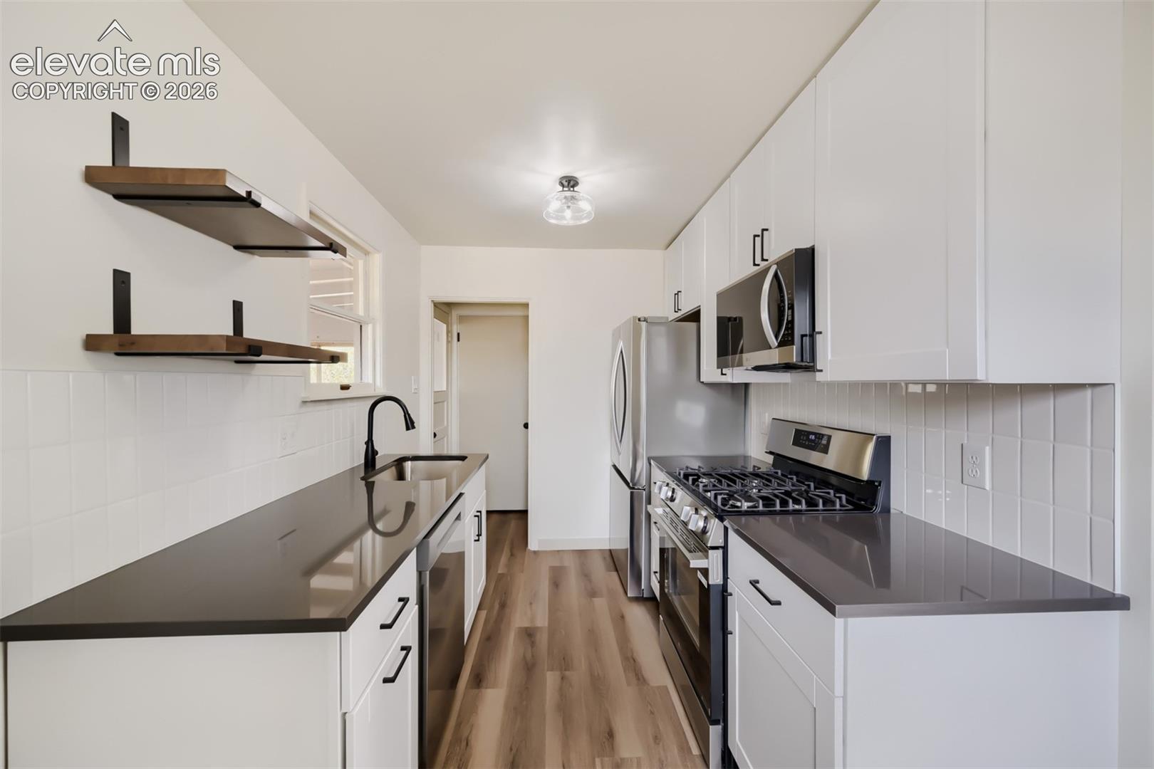 Modern kitchen featuring white cabinetry, dark countertops, wood-finish flooring, stainless steel appliances, and a white tile backsplash