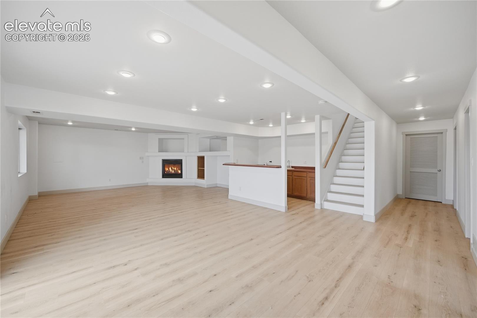 Open-concept interior featuring light wood-finish flooring, recessed lighting, a fireplace with a built-in mantle, a wet bar with cabinetry, and a staircase with a handrail