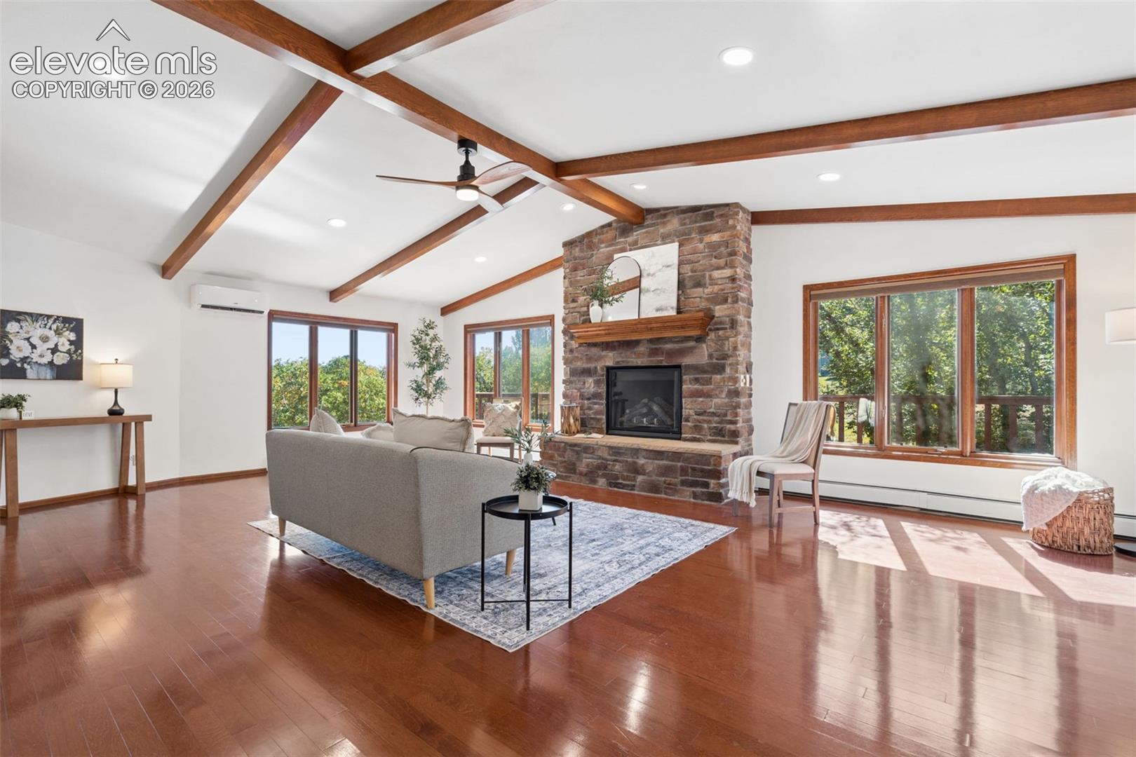 Vaulted ceiling with exposed wood beams, stone-clad fireplace, wood-finish flooring, recessed lighting, and multiple large windows