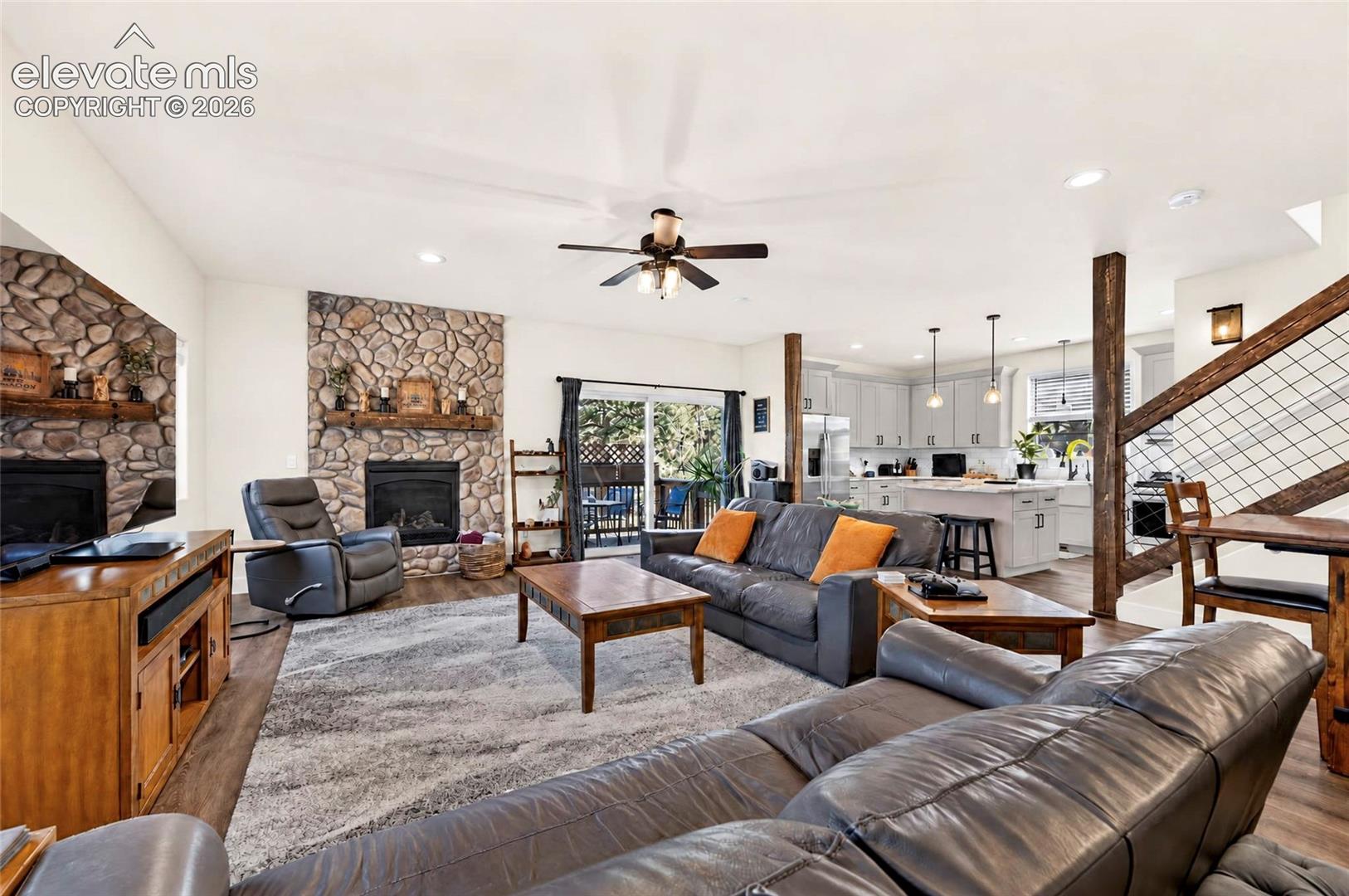 Living area featuring a floor-to-ceiling river rock fireplace with a natural wood mantel, wood-finish flooring, exposed timber support columns, and a staircase with a wire-grid railing