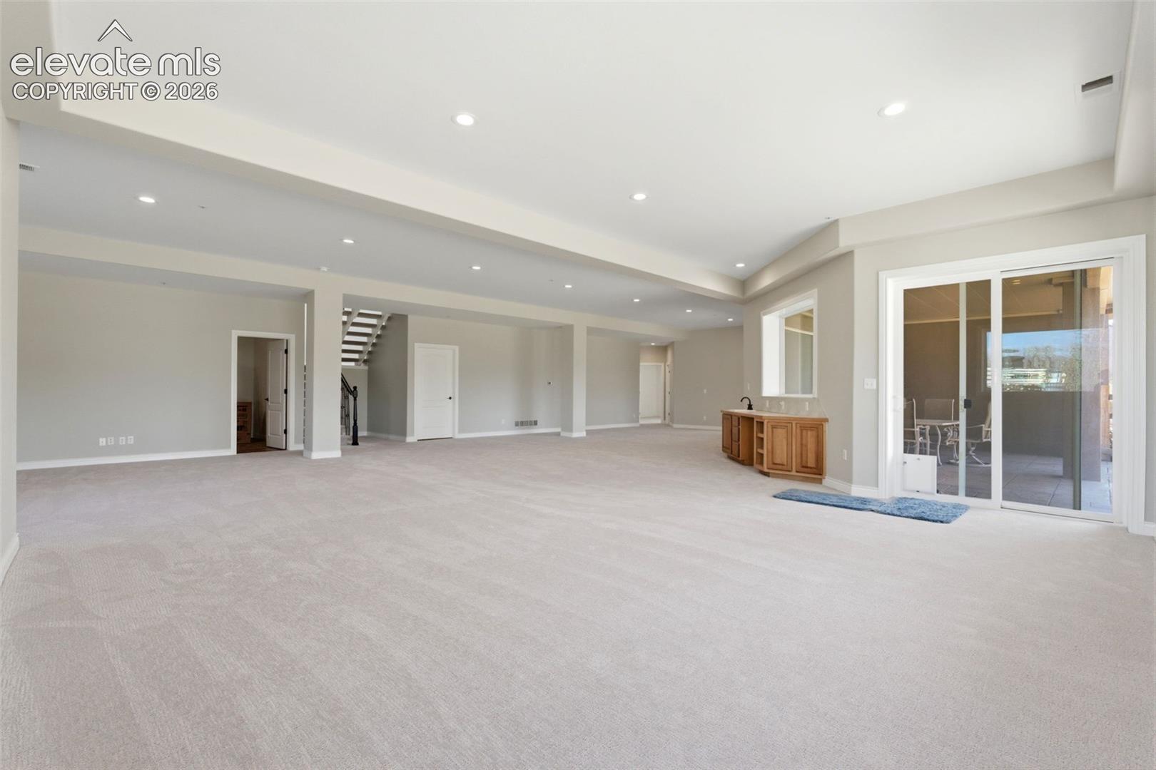 Expansive carpeted area with recessed lighting, built-in wooden cabinetry, sliding glass door access to an outdoor area, and an interior staircase