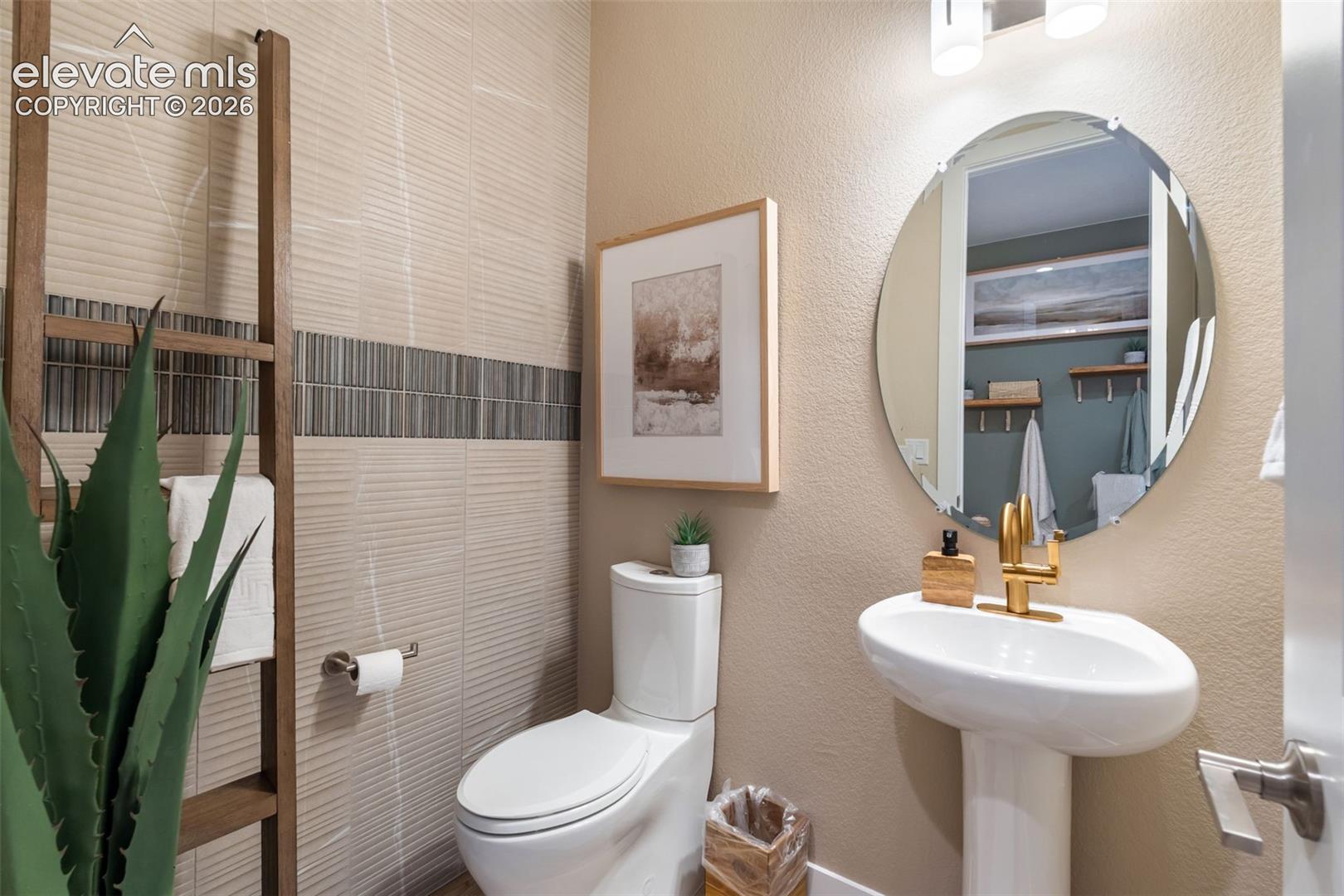 Pedestal sink with a metallic finish faucet, oval wall mirror, textured tiled accent wall, and porcelain toilet