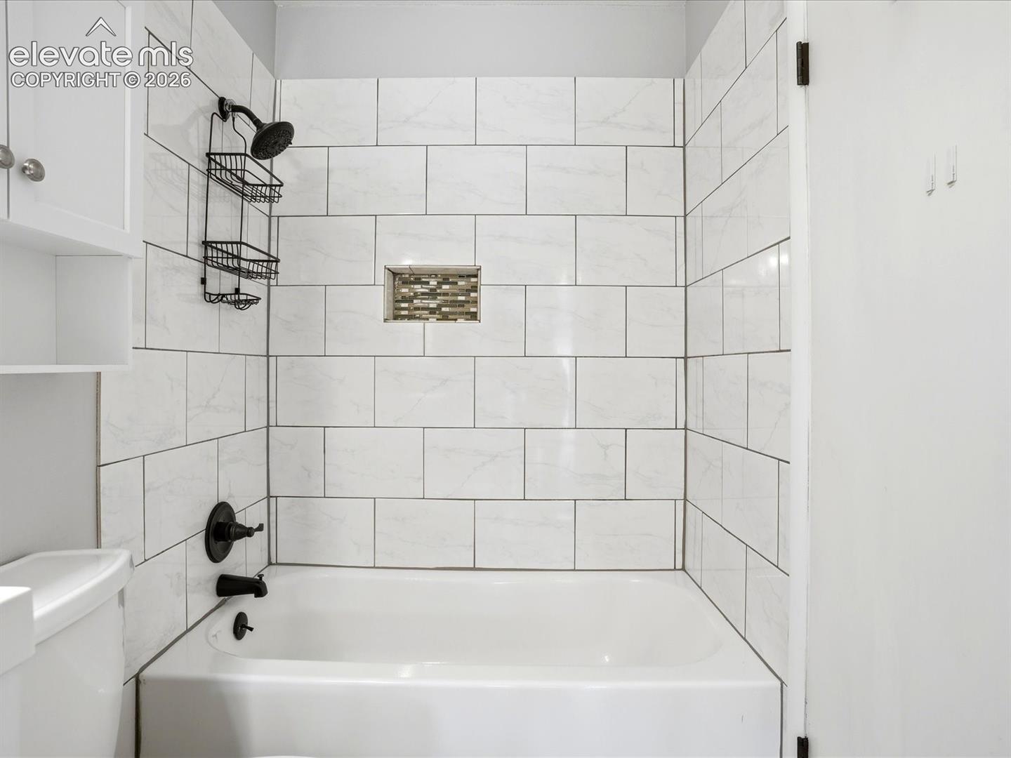Bathtub and shower surround featuring large-format rectangular white tile with subtle grey veining, built-in mosaic tile recessed niche, and matte black plumbing fixtures