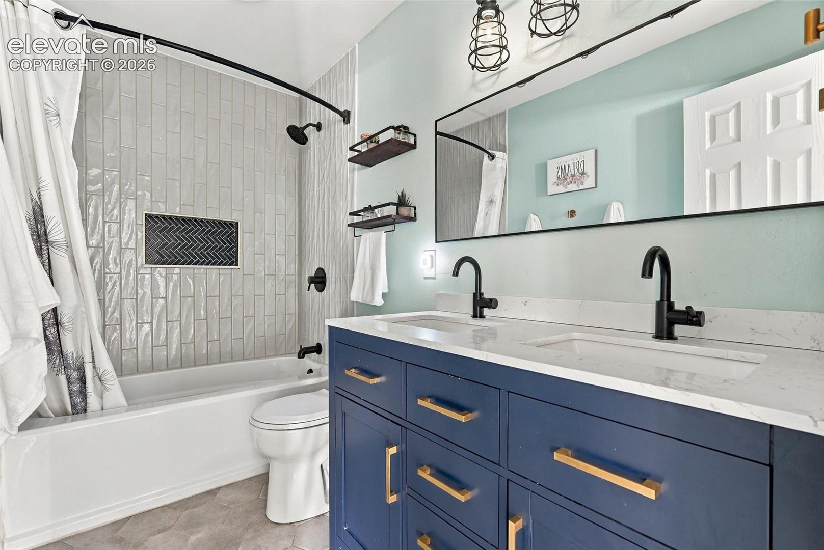 Dual-sink vanity with matte black faucets and a white countertop, blue cabinetry with gold hardware, and a tiled tub surround with a recessed niche