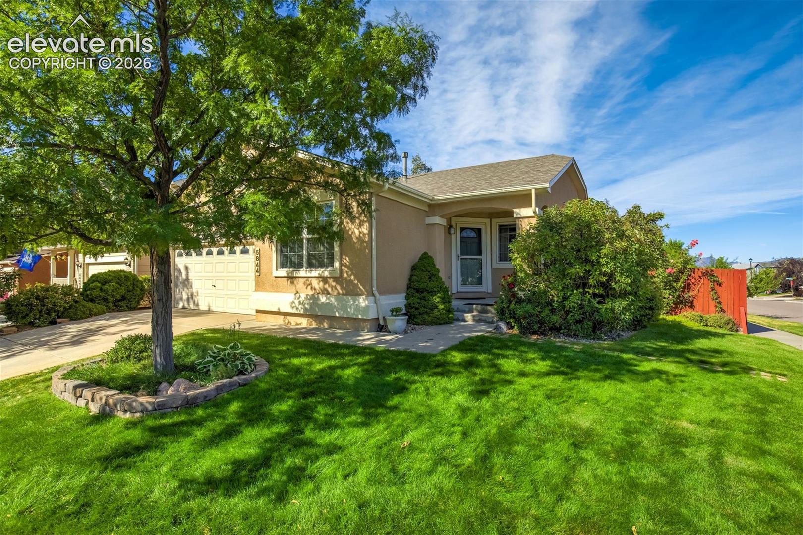 Beige single-story house with an attached garage, shaded by a mature tree and surrounded by a green lawn.