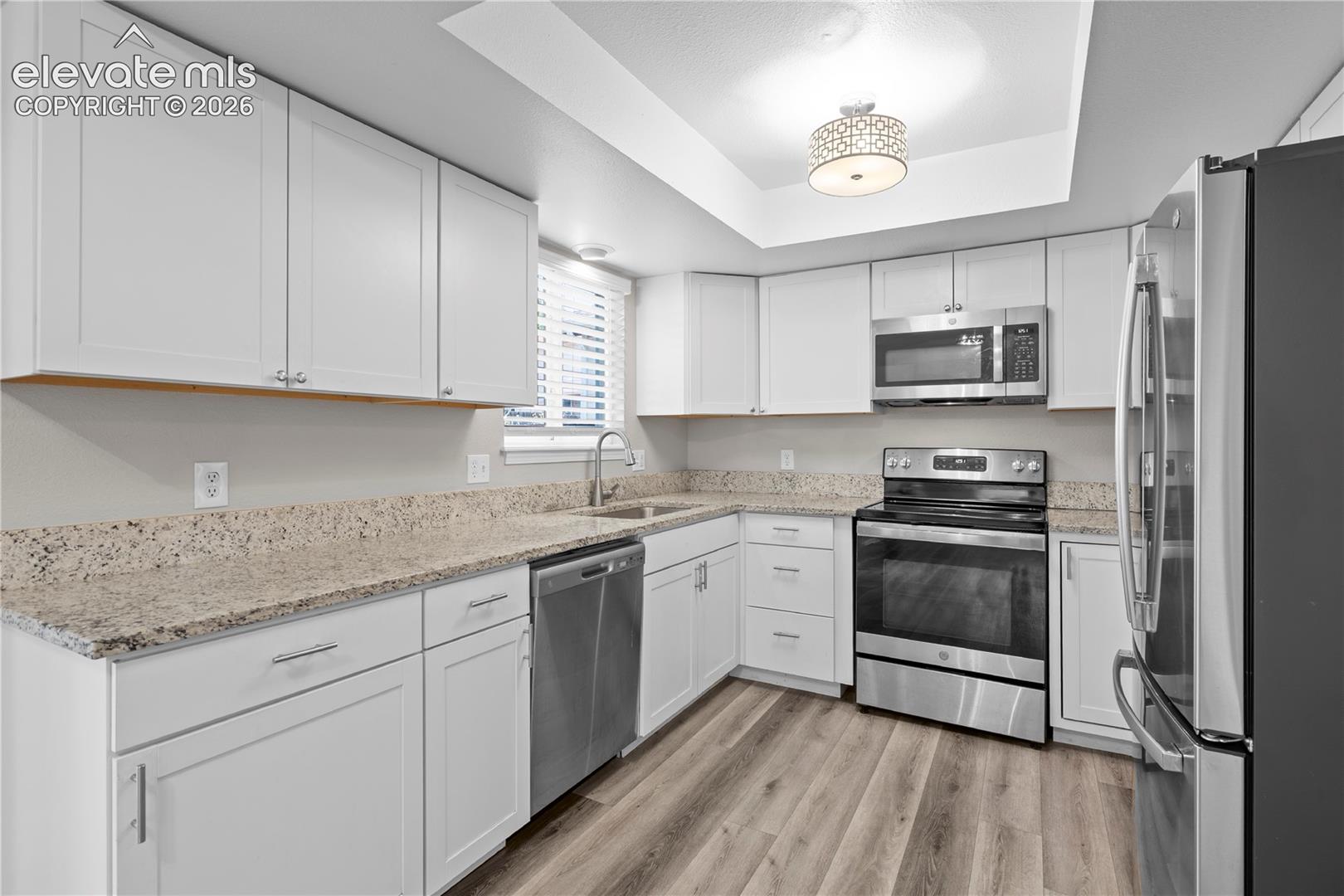 Kitchen featuring white cabinetry, stone-finish countertops, stainless steel appliances, wood-finish flooring, and recessed tray ceiling lighting