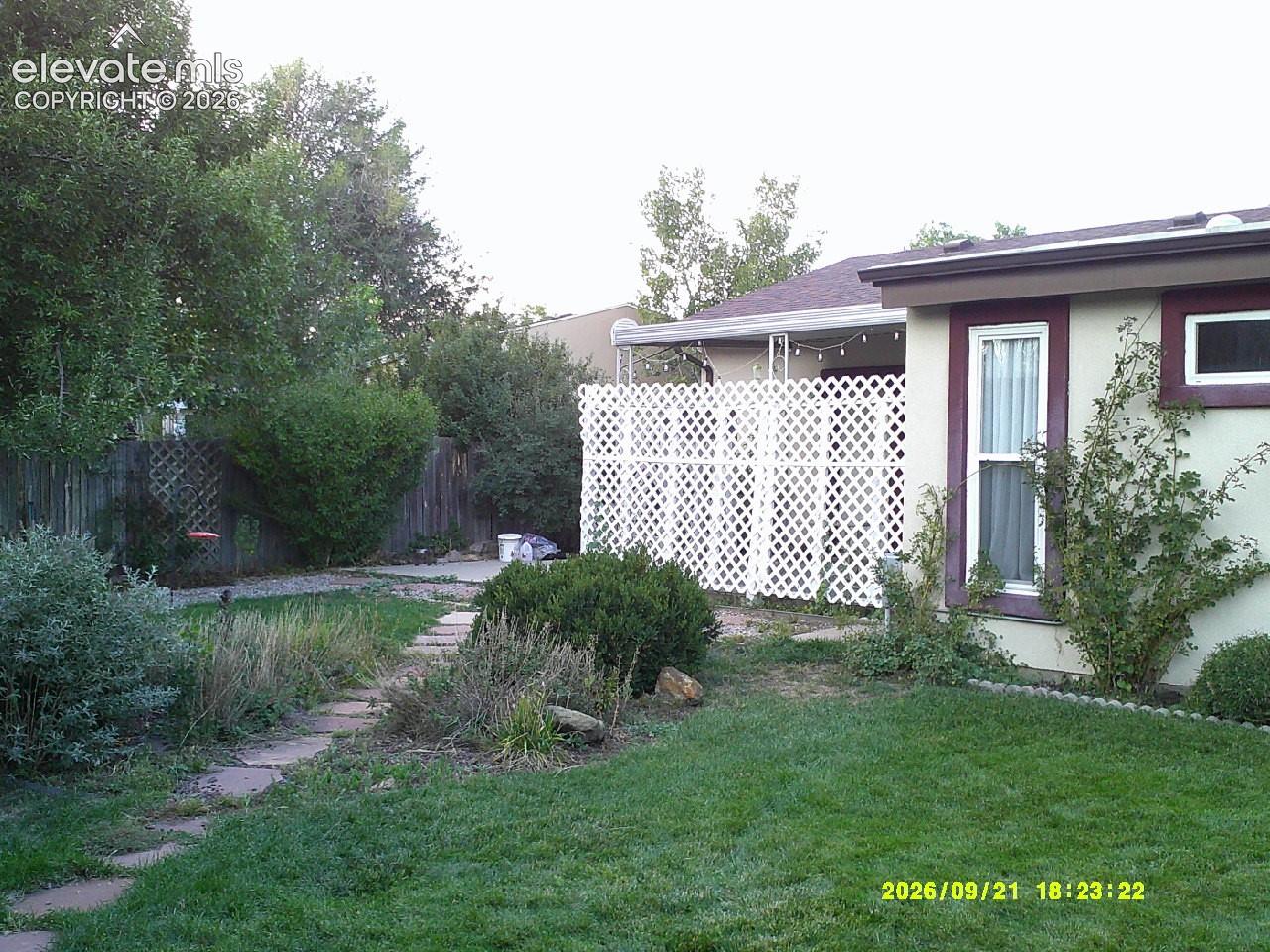 Expansive green lawn, stone-paved pathway, mature trees, white lattice privacy screen, and stucco exterior