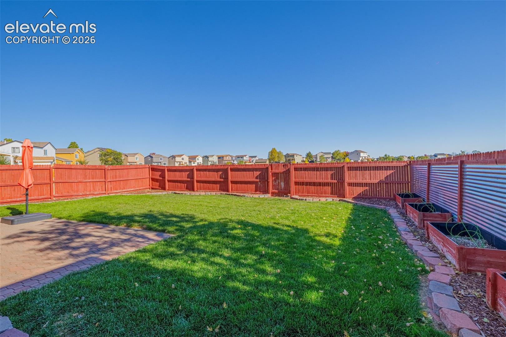 Fenced backyard featuring a paver patio, lawn area, metal-paneled fencing, and elevated planting beds with stone borders
