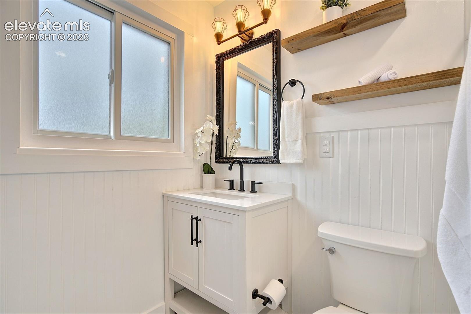 Bathroom featuring a white vanity with a composite countertop, matte black faucet, beadboard wainscoting, and natural wood-finish shelving