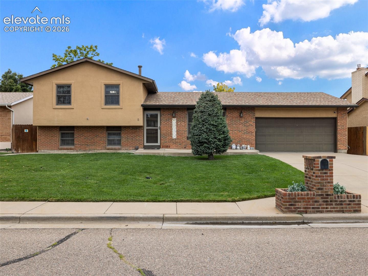 Brick and stucco exterior, attached garage, well-maintained lawn, paved driveway, and a brick mailbox planter