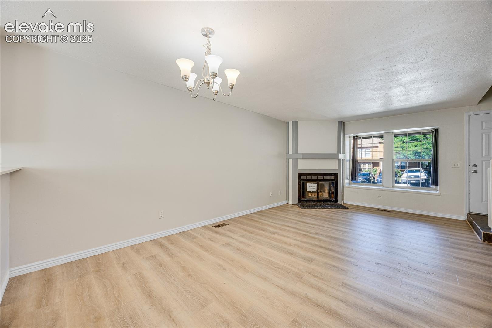 Spacious room featuring wood-finish flooring, a built-in fireplace with a hearth, a large window, a decorative chandelier, and a white entry door