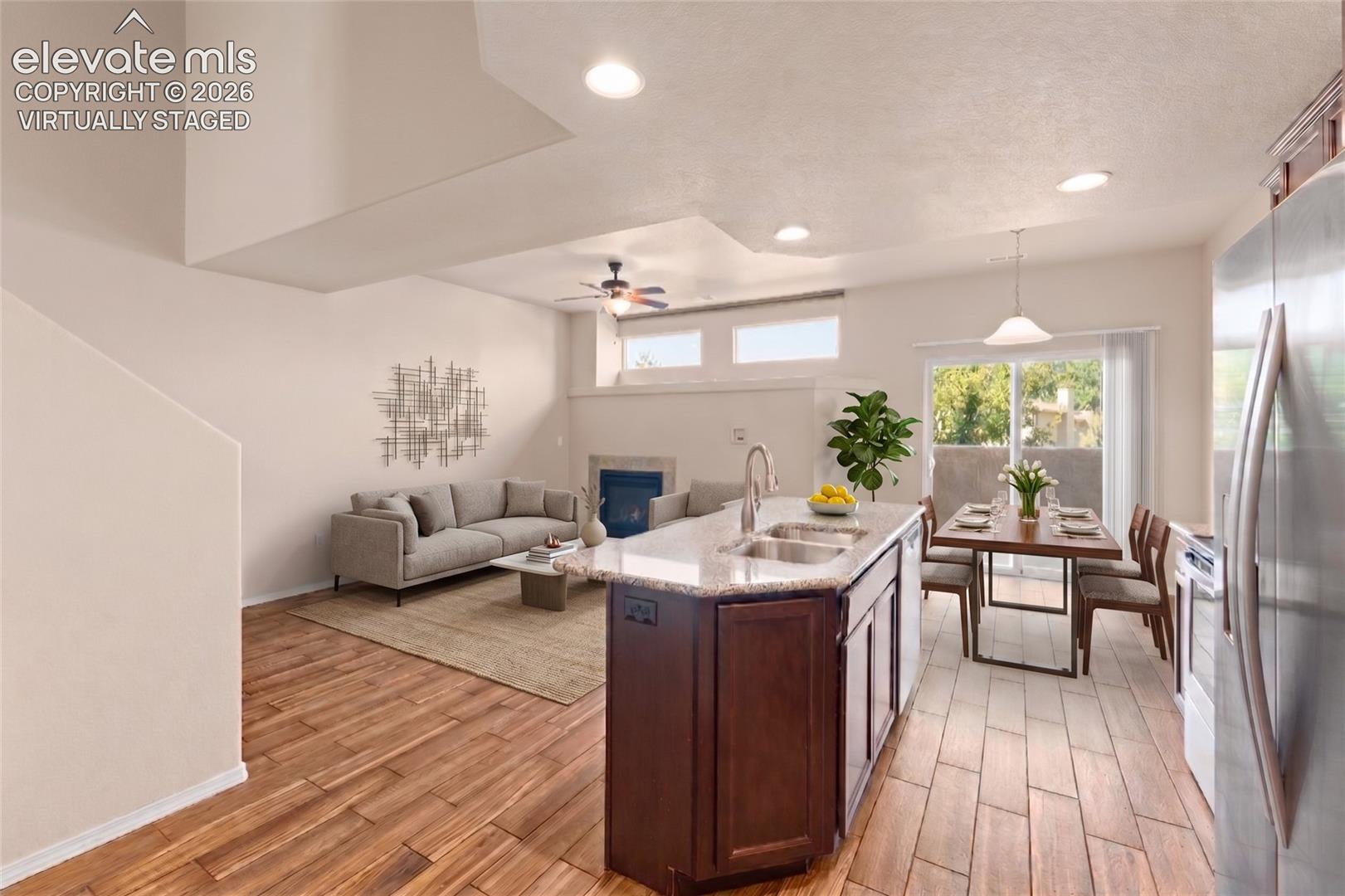 Open-concept living area featuring wood-finish flooring, a kitchen island with dual sinks, a dining area with a sliding glass door, and a living room with an integrated fireplace - virtually staged