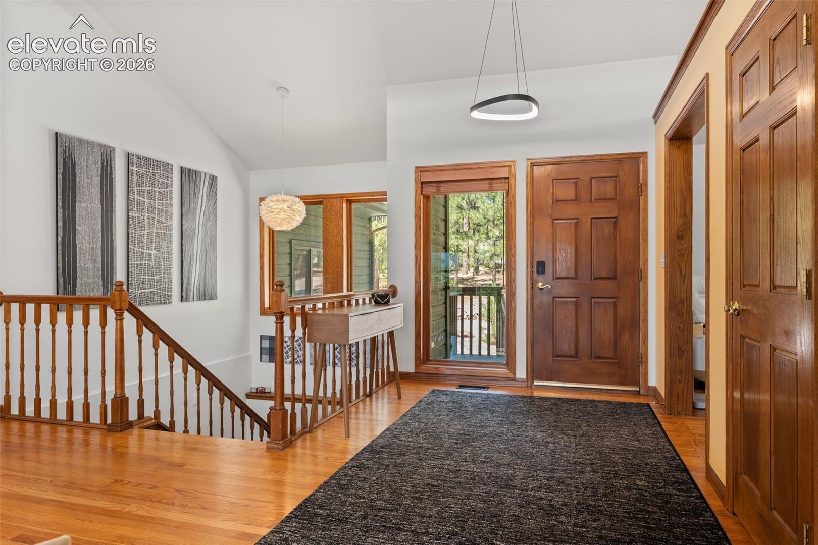 Warm wood-finish flooring extends through this welcoming entry, featuring a solid wood door with sidelight window, a vaulted ceiling, and wood banister staircase