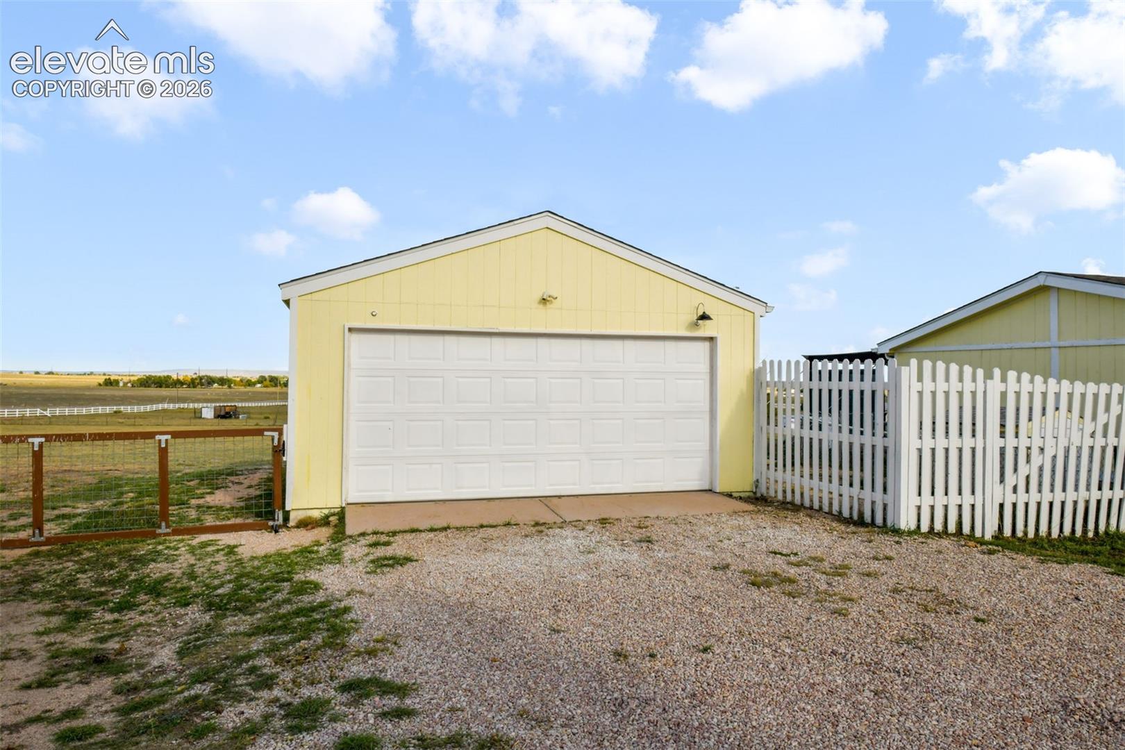 Detached garage with a white panel door, exterior lighting, and a gravel driveway
