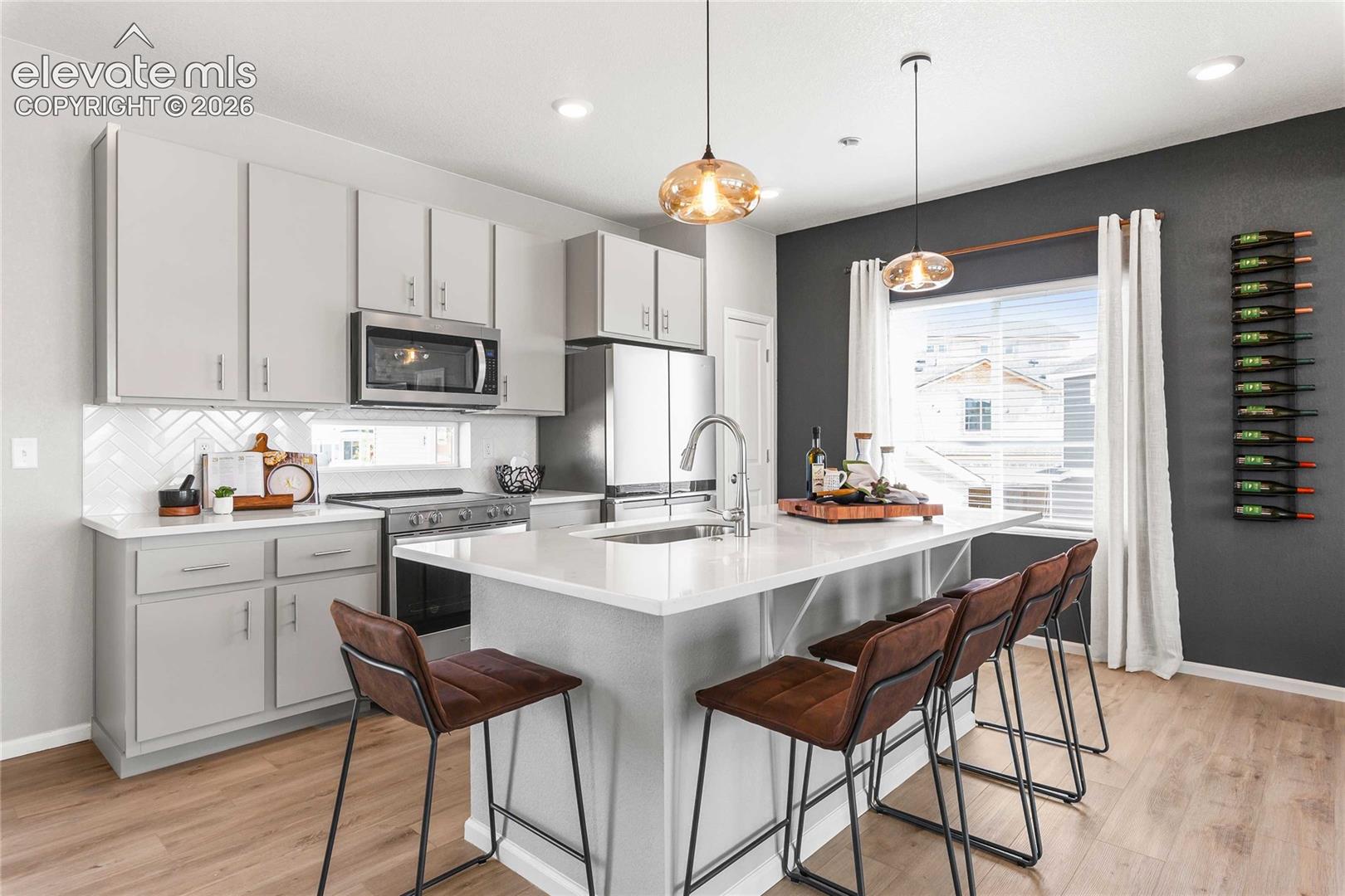 Modern kitchen featuring soft-close cabinetry with brushed nickel hardware, quartz waterfall-edge island countertop, stainless steel appliances including a built-in microwave, chrome pull-down faucet, pendant lighting, wood-finish flooring, and an accent wall in charcoal gray with integrated wine storage
*photos of a model home*