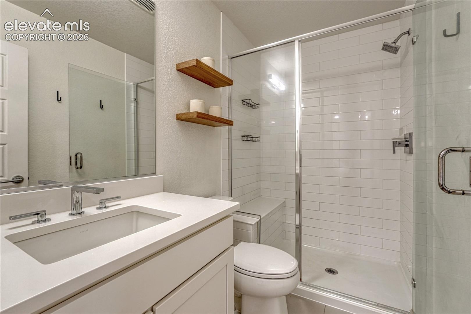 Bathroom featuring a white vanity with a solid-surface countertop, rectangular undermount sink, glass-enclosed shower with subway tile surround, and wall-mounted wood shelving