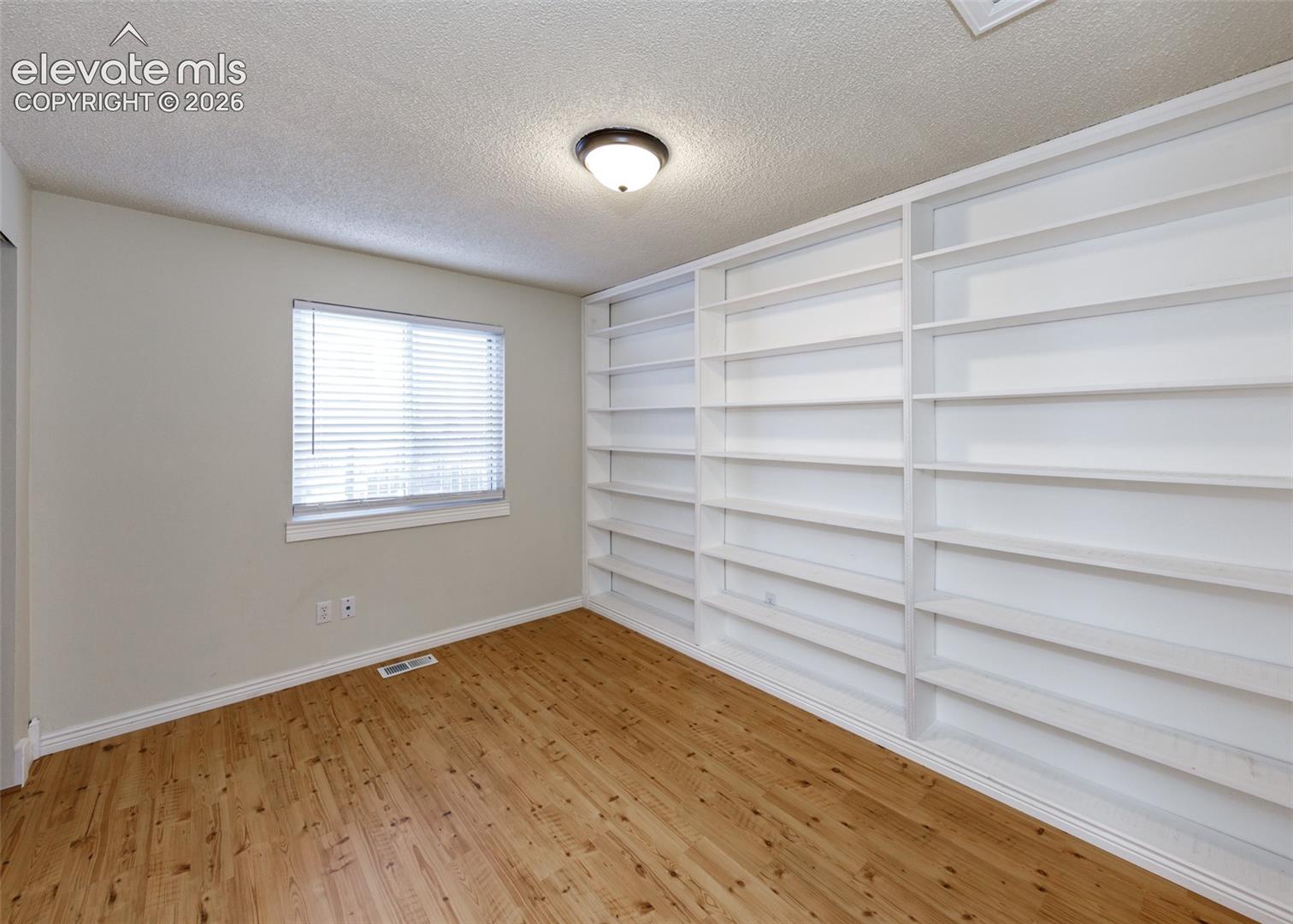 Room featuring wood-finish flooring, built-in floor-to-ceiling shelving, and a dual-pane window with adjustable blinds