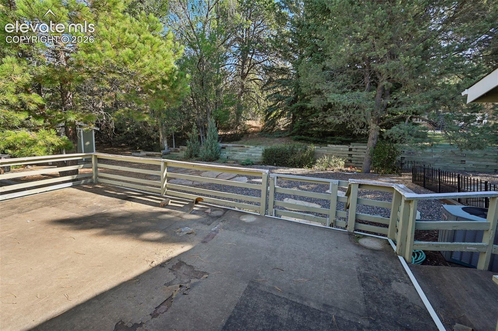 Concrete deck featuring a perimeter wood railing, tiered landscape wall, and established conifer trees