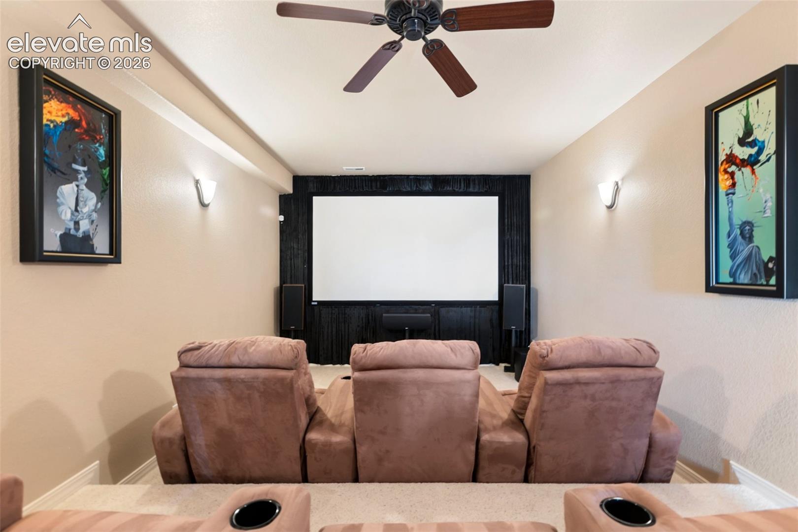 Dedicated media room featuring a large projection screen, dark wood-paneled accent wall, stepped platform flooring, dual wall-mounted sconces, and a ceiling-mounted fan