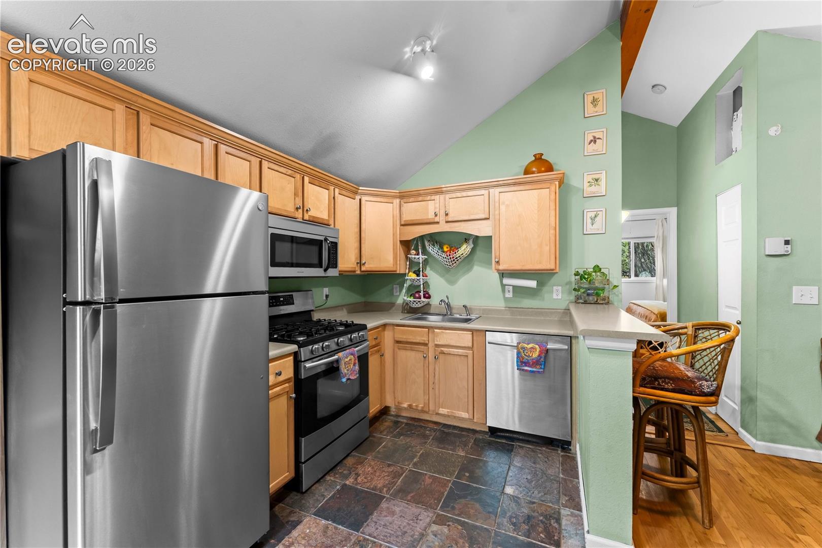 Kitchen featuring wood cabinetry, stainless steel appliances, slate-finish floor tile, and a vaulted ceiling
