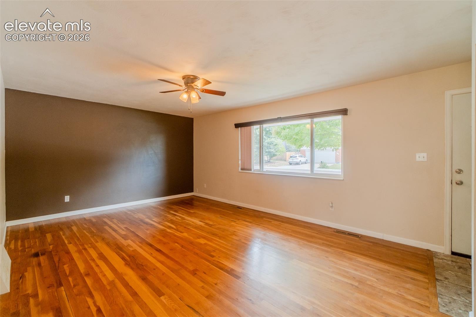 Hardwood flooring, ceiling fan with integrated lighting, large window with natural light, neutral wall colors, and a white entry door