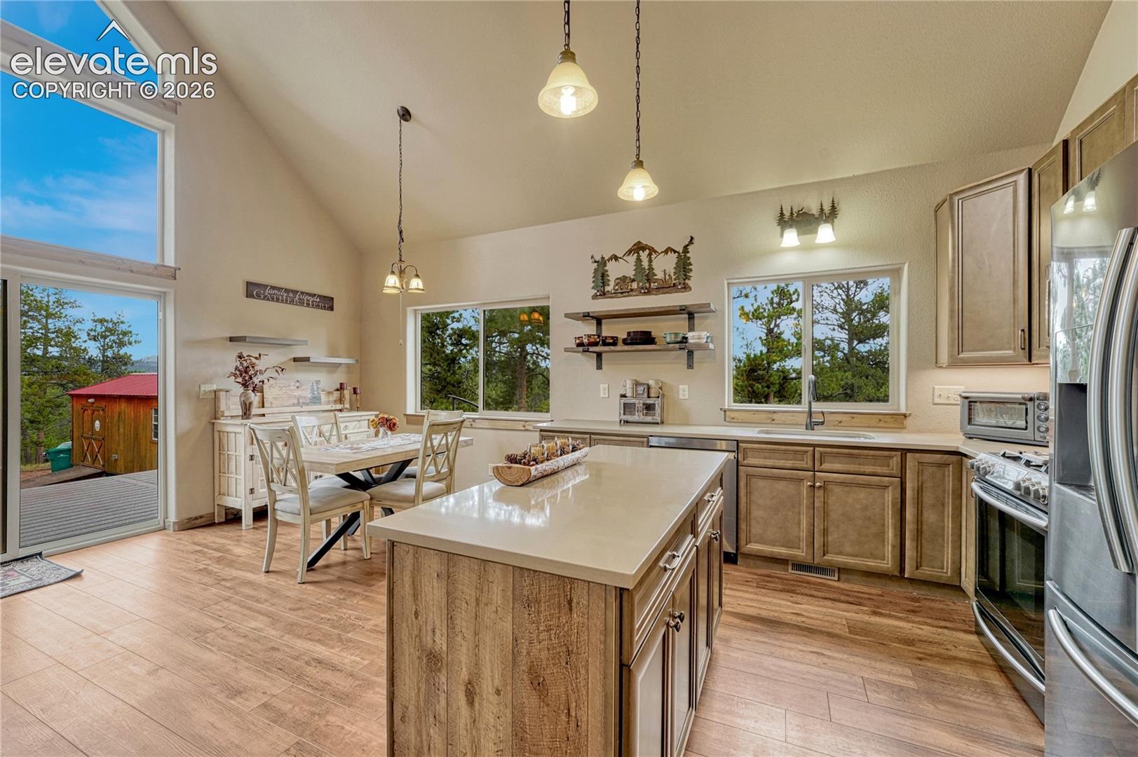 Vaulted ceilings featuring pendant lighting, expansive windows, kitchen island with stone-finish countertop, wood-finish flooring, and cabinetry with stainless steel appliances