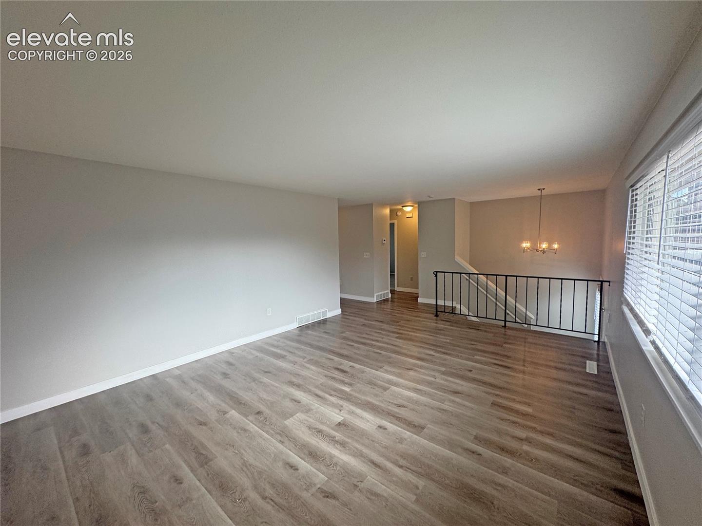 Open-concept living space featuring wood-finish flooring, neutral wall paint, horizontal window blinds, and a metal staircase railing